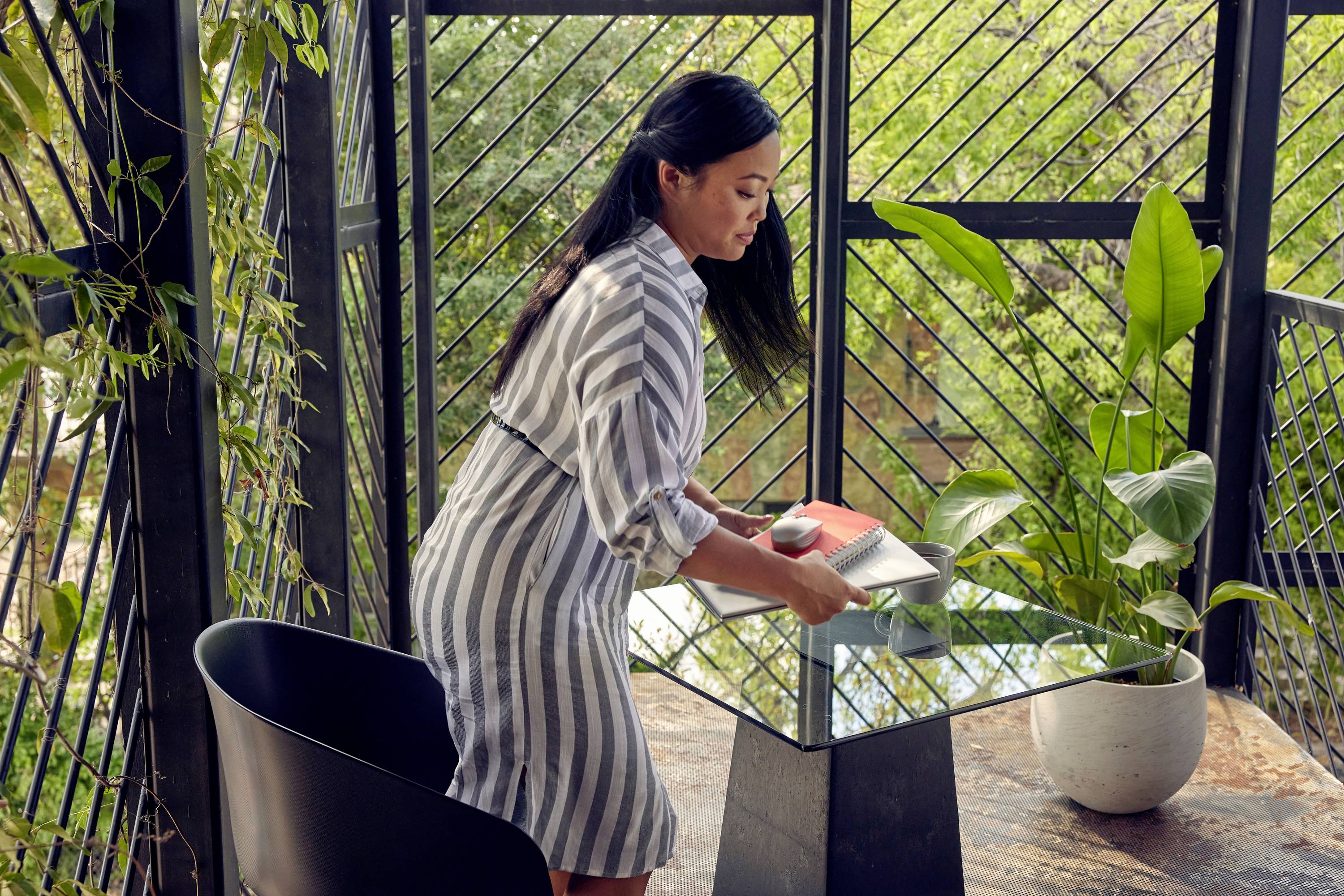 Frau in gestreiftem Kleid räumt einen Glastisch auf einer Terrasse auf. Die Umgebung ist von Pflanzen umgeben und es ist sonnig.