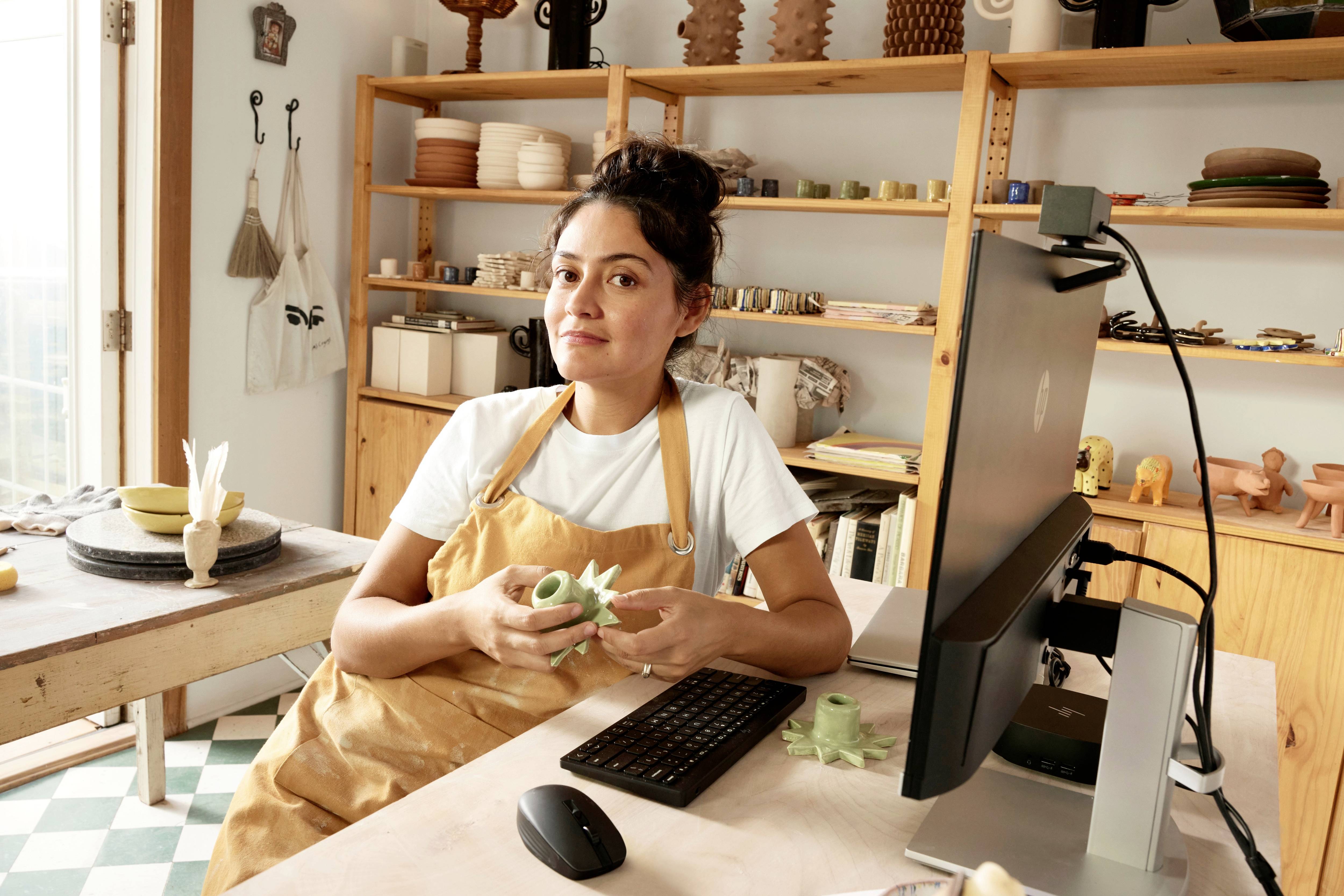 Eine Person in Arbeitskleidung sitzt am Schreibtisch in einem hellen Raum und hält Tonobjekte. Im Hintergrund sind Regale mit Keramik.