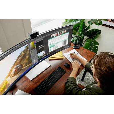 Person arbeitet an einem Schreibtisch mit einem großen, gebogenen Monitor, schreibt Notizen, während mehrere Programmfenster offen sind.