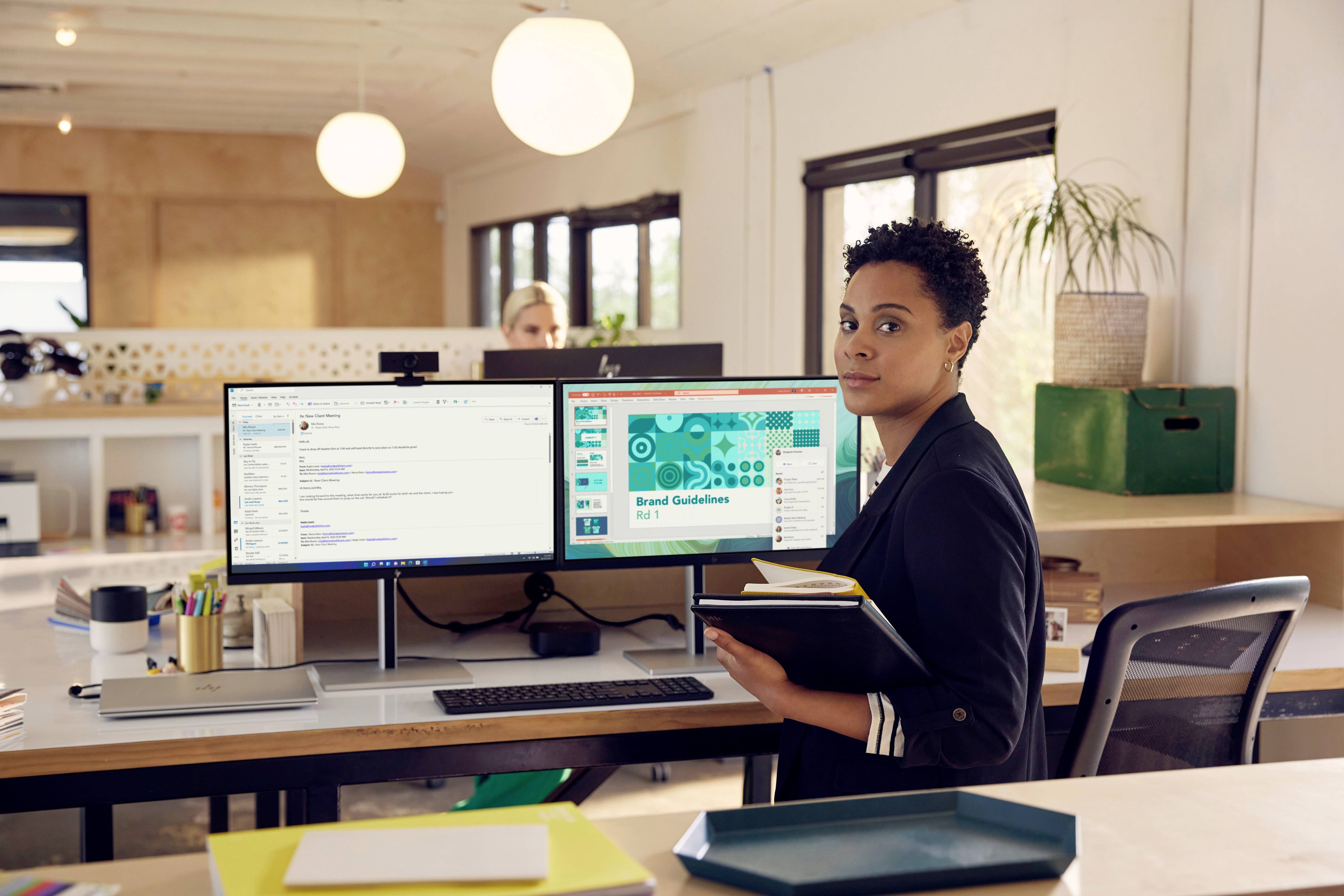 Eine Person sitzt an einem Schreibtisch mit zwei Monitoren, auf denen E-Mails und Markentrichtlinien zu sehen sind. Sie hält ein Notizbuch.