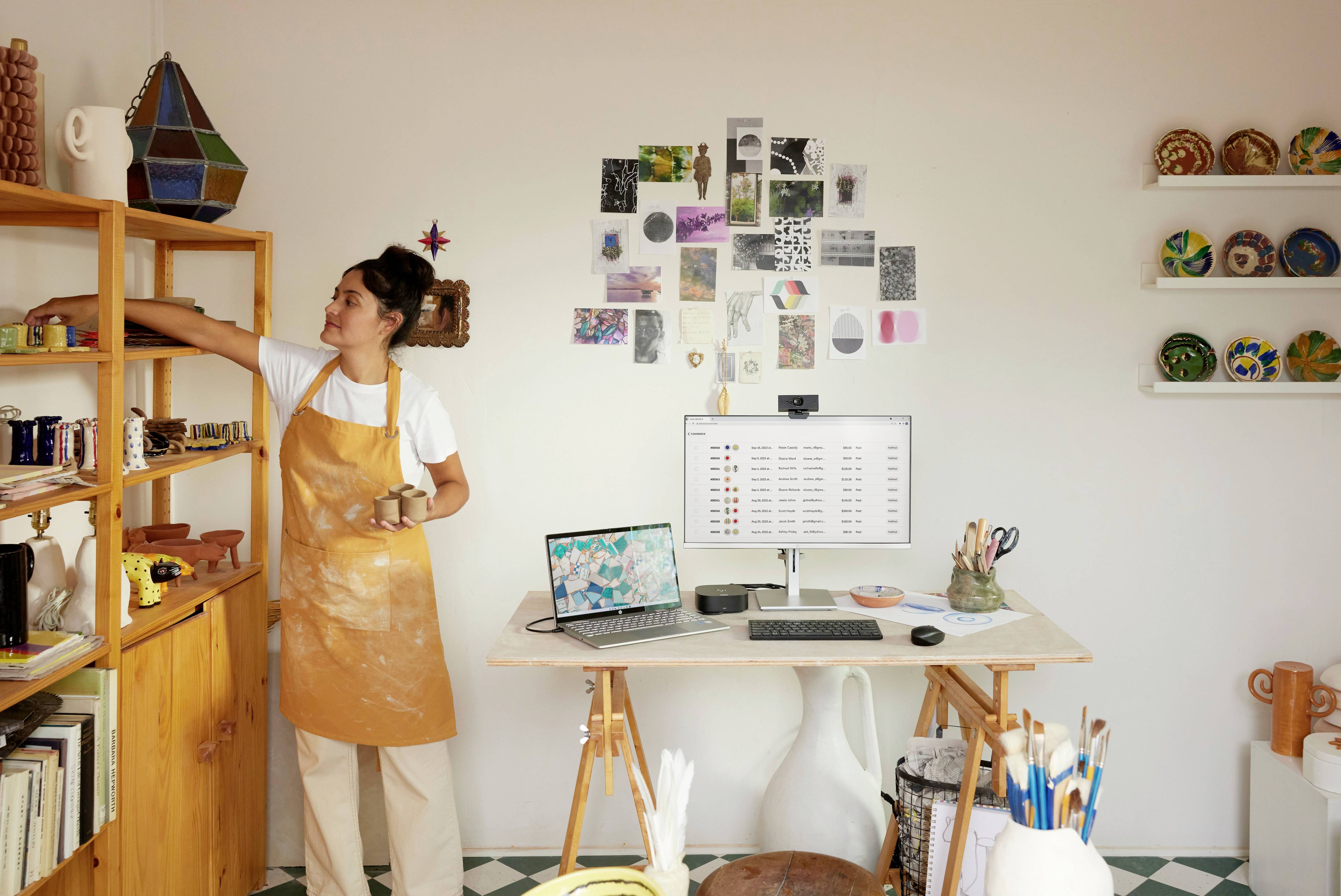 Eine Person steht in einem Atelier und greift nach Materialien im Regal. Vor ihr steht ein Schreibtisch mit Computer und Laptop.