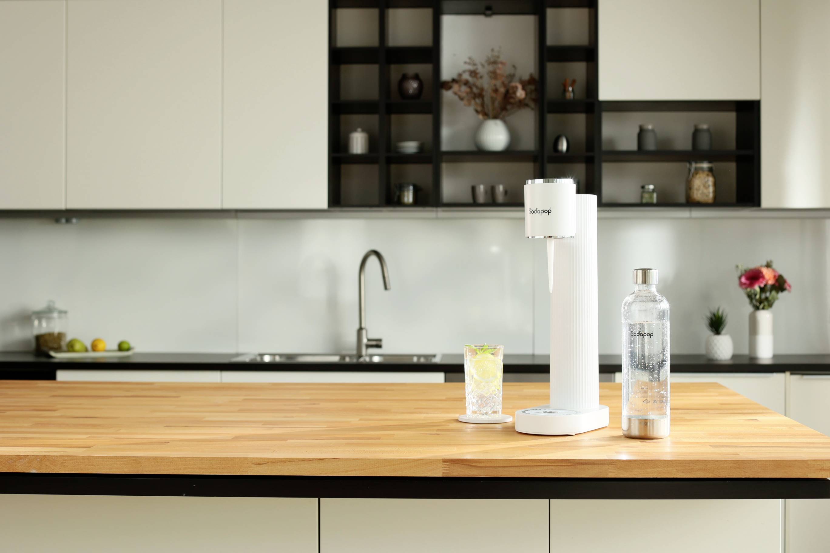 Ein Wassersprudler steht auf einer Holzplatte in einer modernen Küche. Daneben ein Glas mit Sprudelwasser und eine Karaffe.