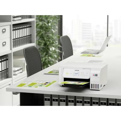 Ein Drucker druckt Broschüren auf einem Bürotisch. Im Hintergrund sind ein Laptop, Ordnerregale und Pflanzen zu sehen.
