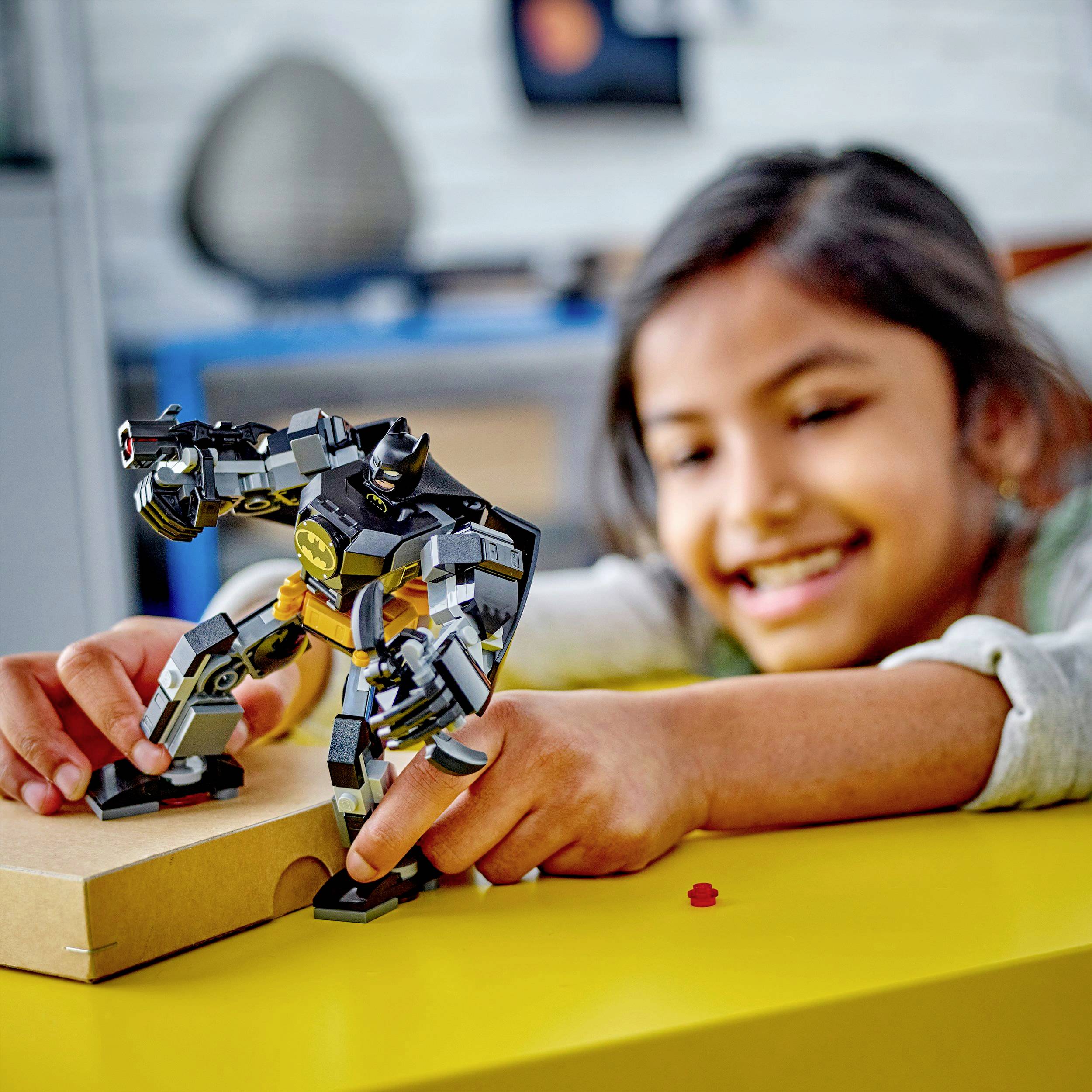 Ein Kind spielt lächelnd mit einer schwarzen Roboterfigur auf einem gelben Tisch. Im Hintergrund ist ein unscharfer Raum sichtbar.