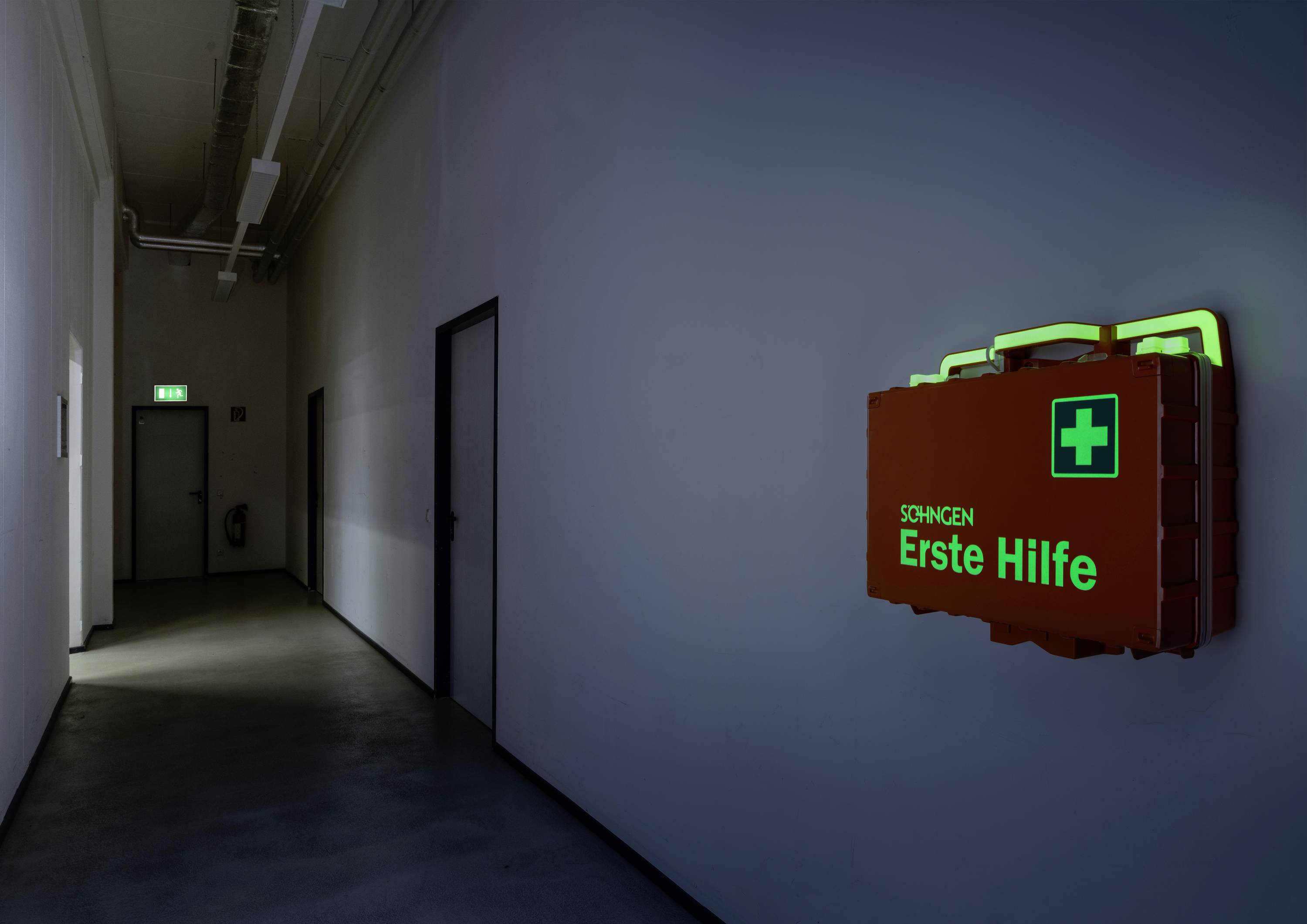 Ein roter Erste-Hilfe-Kasten mit grünem Kreuz und 'Erste Hilfe'-Text hängt an einer dunklen Flurwand, beleuchtet durch Leuchtschrift.