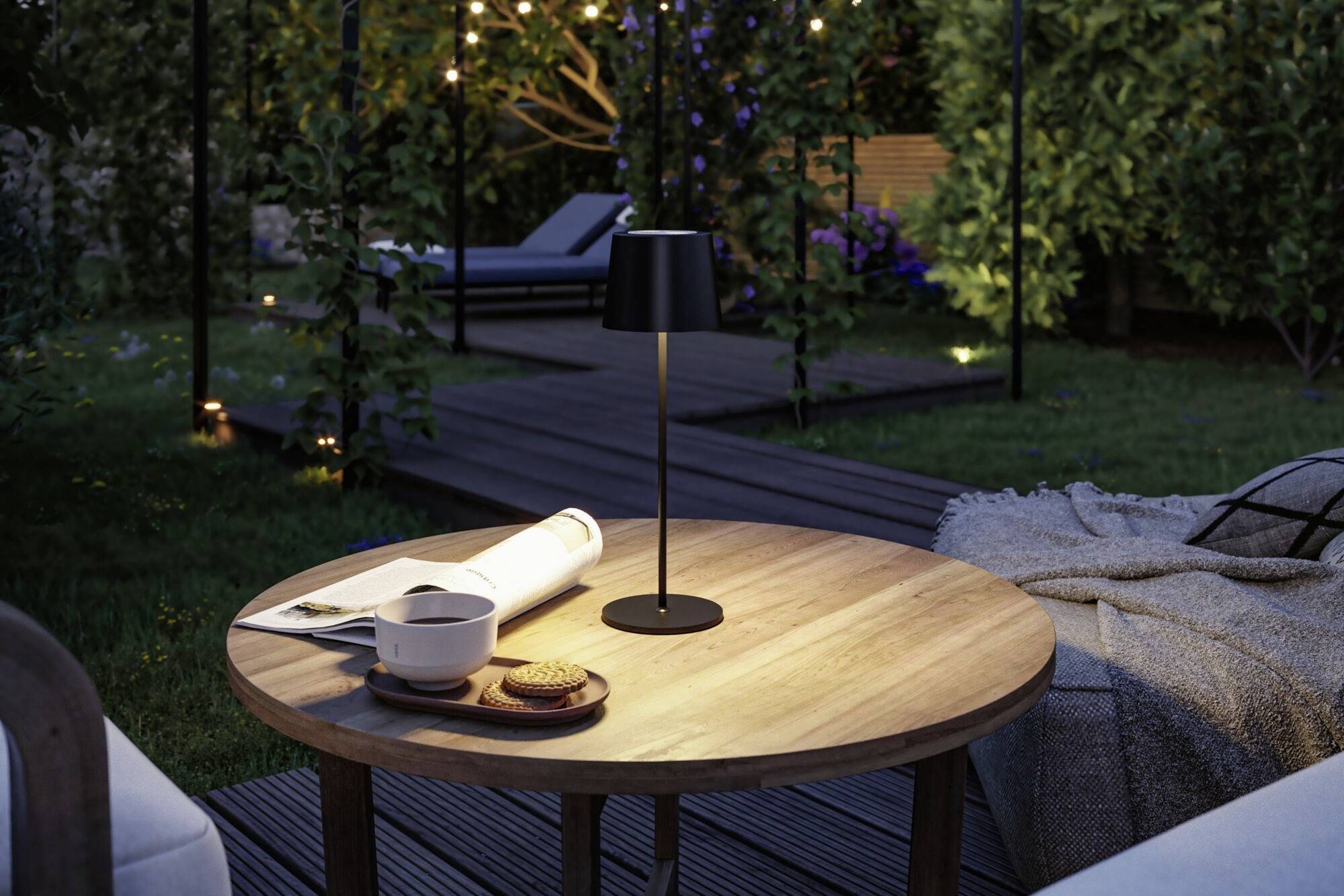 Ein runder Holztisch im Garten mit Buch, Tasse und Keksen darauf. Im Hintergrund Abendstimmung mit Lichterketten, Liegen und Pflanzen.