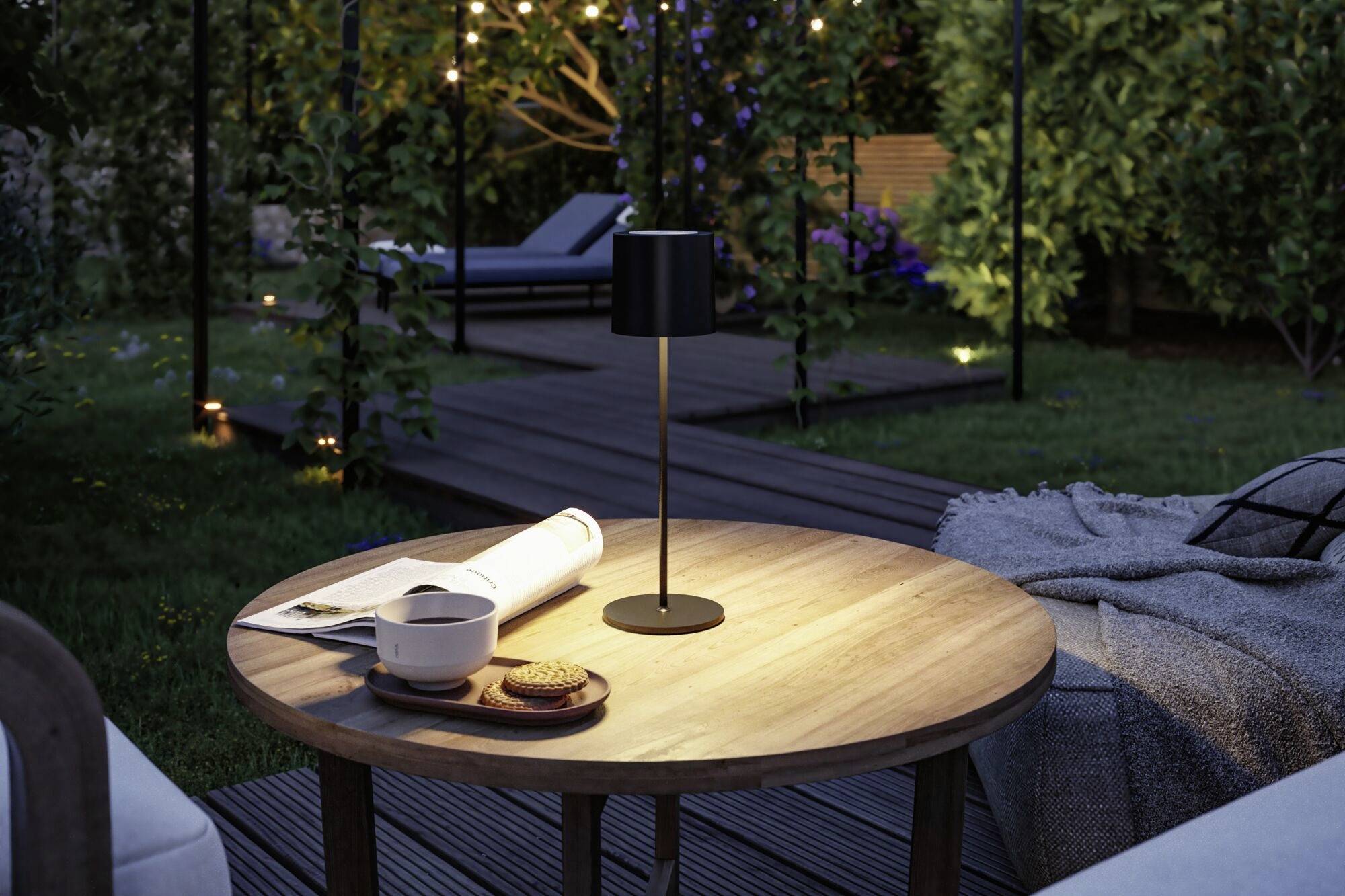 Runder Holztisch im Garten bei Abenddämmerung mit Buch, Kaffeetasse und Keksen. Hintergrund zeigt blühende Pflanzen und Beleuchtung.