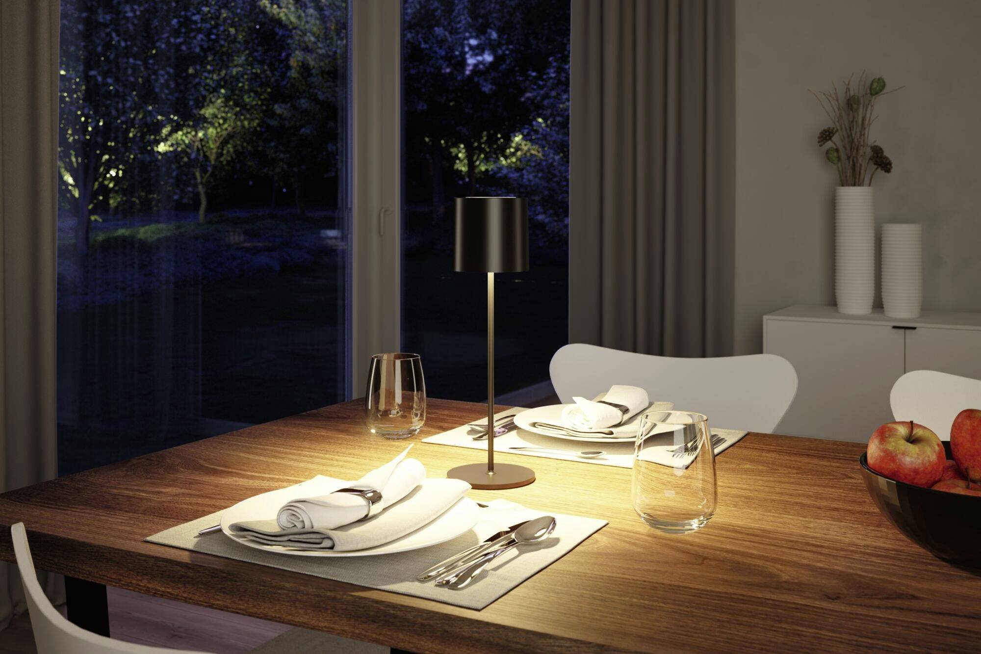 Ein Esszimmertisch ist für zwei Personen gedeckt, mit Besteck, Gläsern und Stoffservietten. Eine Lampe beleuchtet den Tisch. Im Hintergrund sind Fenster mit Blick in einen Garten.