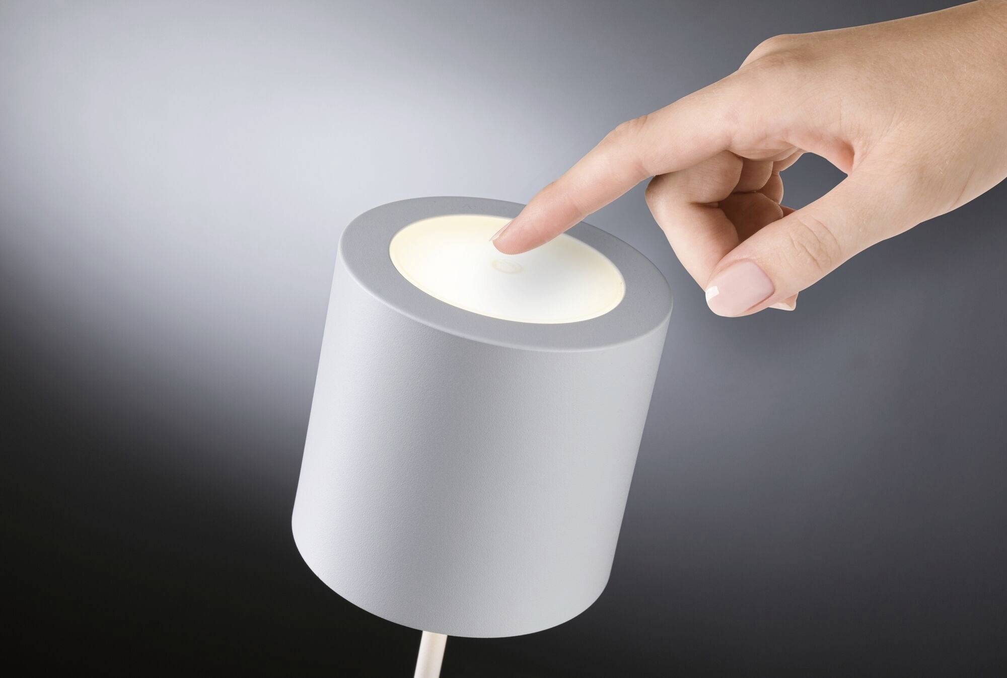 Eine Hand berührt leicht den Schalter einer modernen, zylindrischen Tischlampe mit grauem Gehäuse, die eingeschaltet ist.