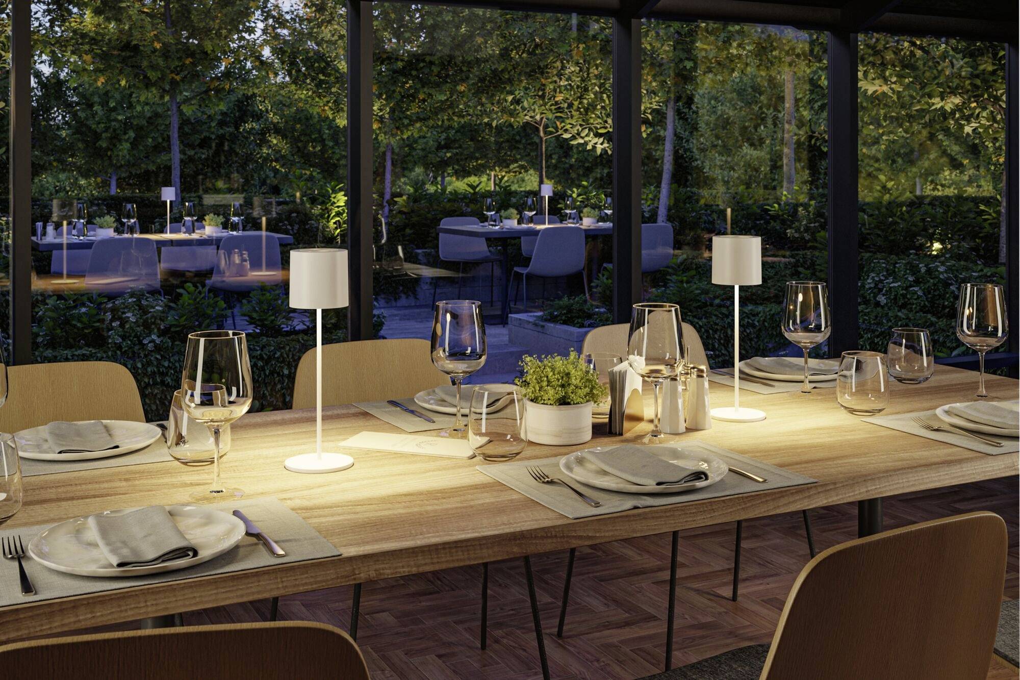 Restauranttisch für sechs Personen, gedeckt mit Weingläsern, Tellern und Besteck, sanft beleuchtet; Gartenbereich im Hintergrund.