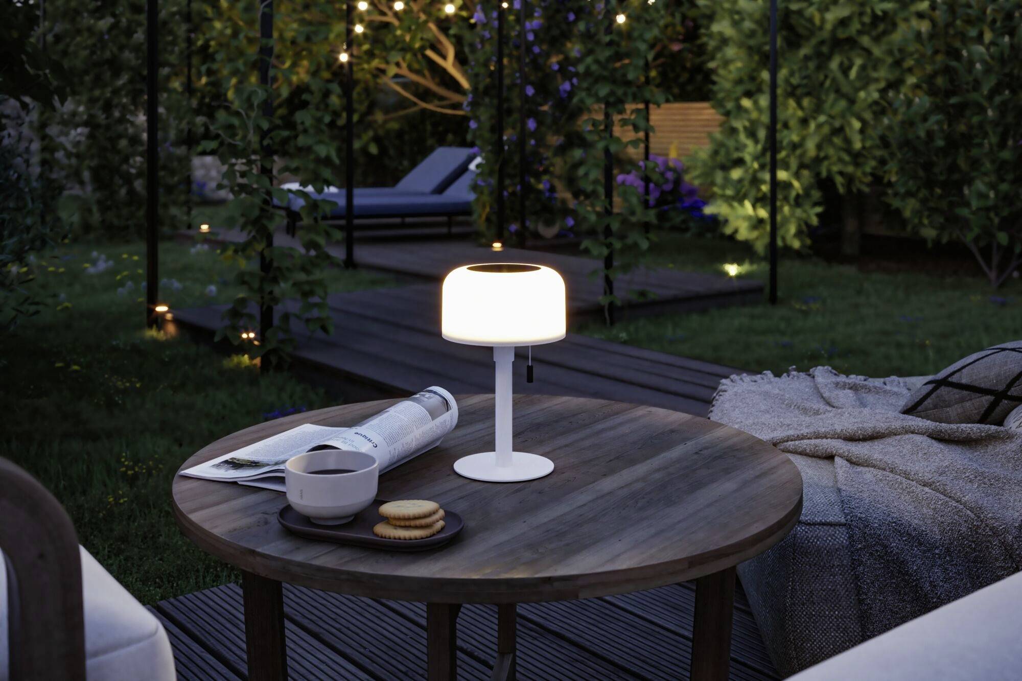 Ein runder Holztisch im Garten bei Dämmerung, mit einer kleinen Lampe, eine Tasse Kaffee und Kekse darauf, umgeben von Pflanzen.