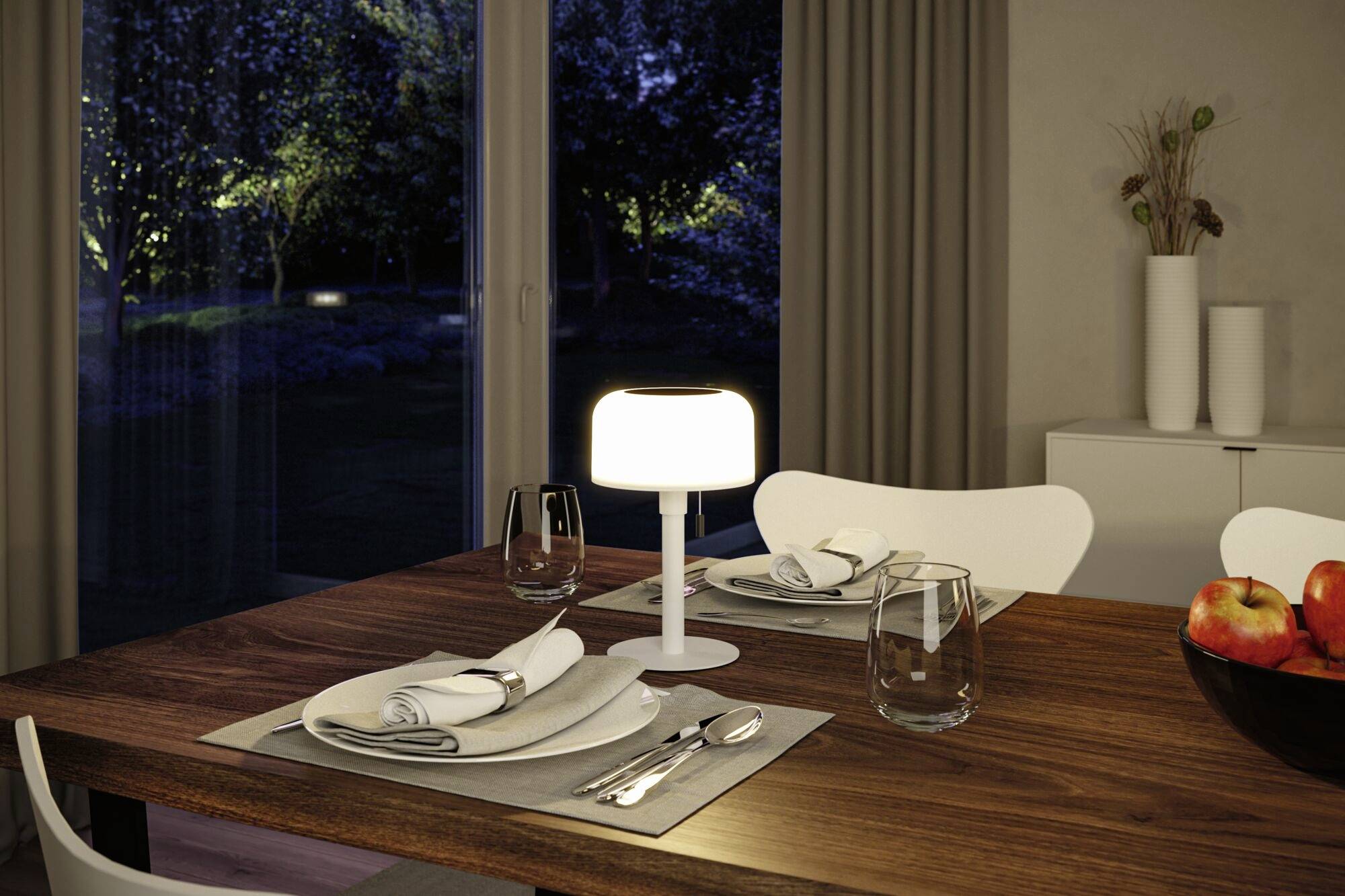 Der Esszimmertisch ist für zwei Personen gedeckt, umgeben von gedämmtem Licht durch eine Tischlampe, mit Blick auf den Garten im Hintergrund.