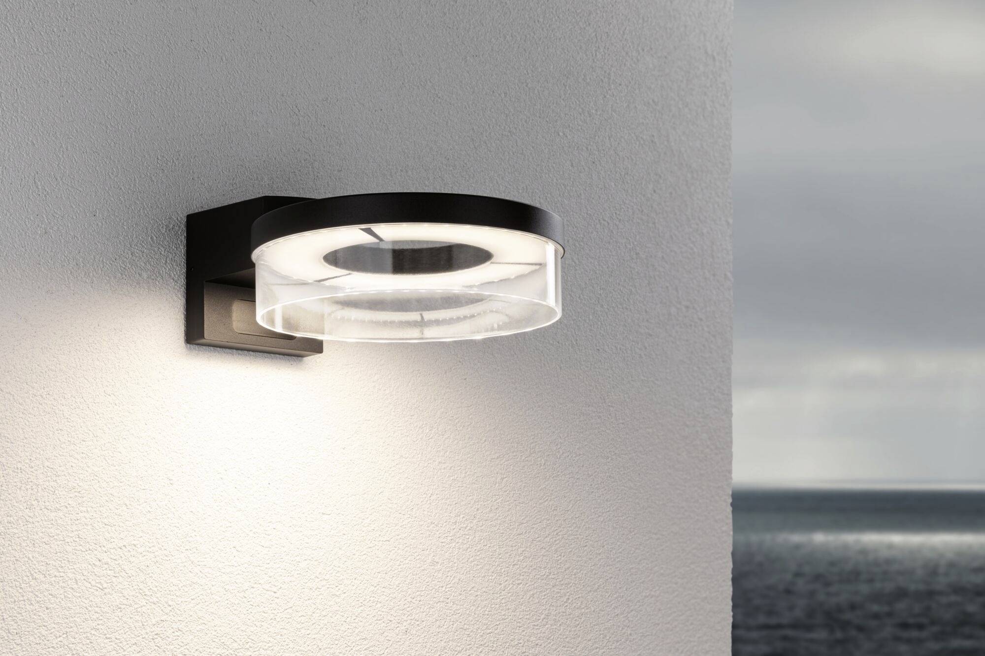 Eine moderne Außenwandleuchte beleuchtet eine weiße Wand. Im Hintergrund ist ein bewölkter Himmel über dem Meer zu sehen.