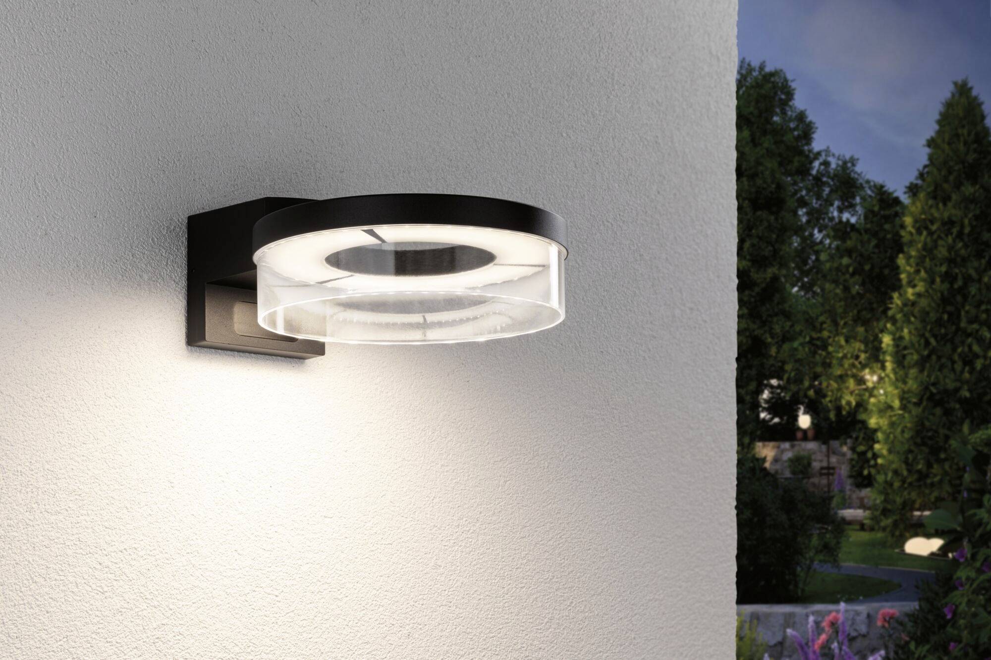 Außenwandlampe mit modernem Design leuchtet sanft in der Dämmerung; montiert an weißer Wand, Pflanzen und Garten im Hintergrund.