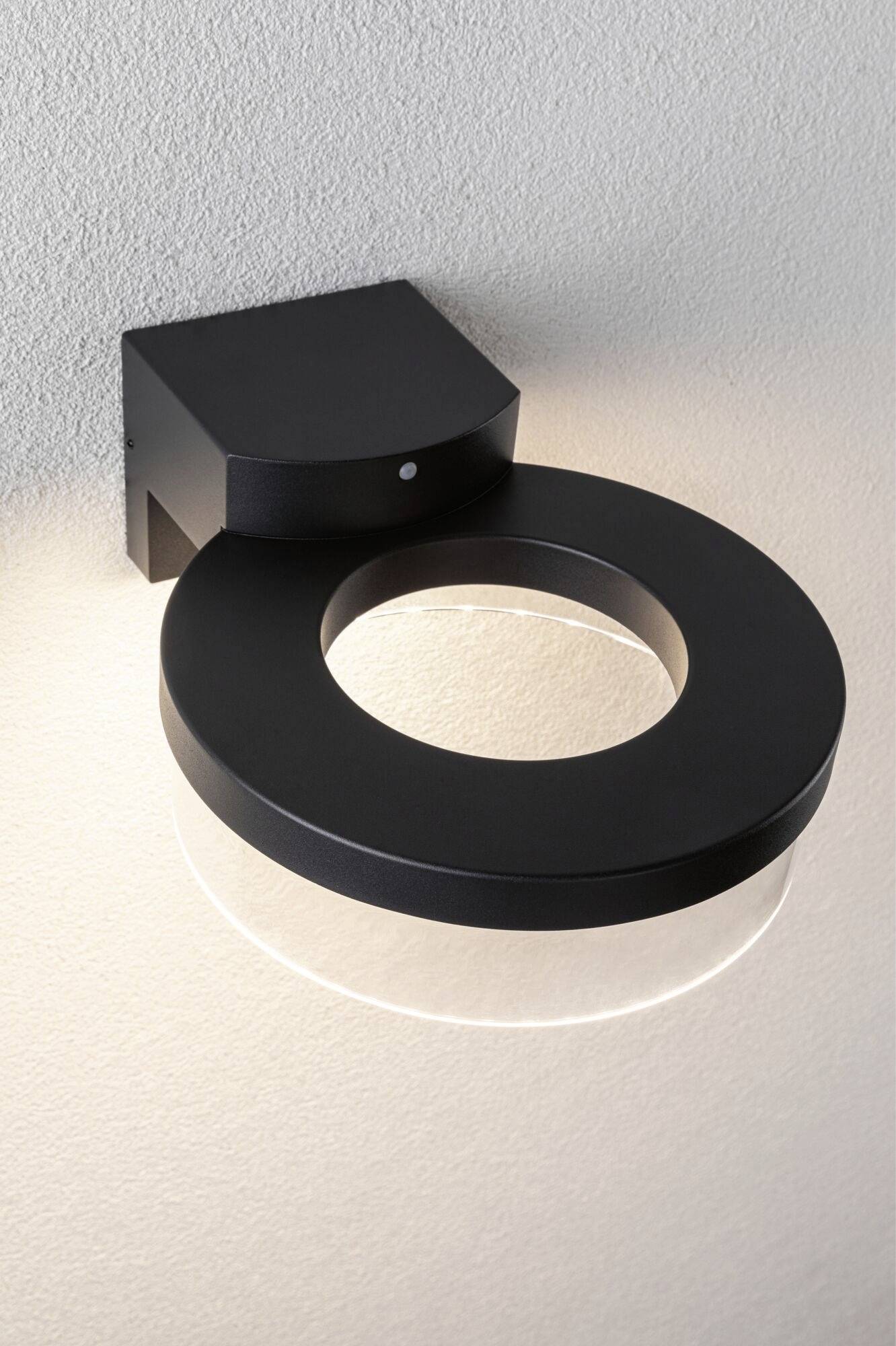 Schwarze, ringförmige Wandleuchte an einer hellen Wand, strahlt weiches Licht nach unten und erzeugt einen gleichmäßigen Lichteffekt.