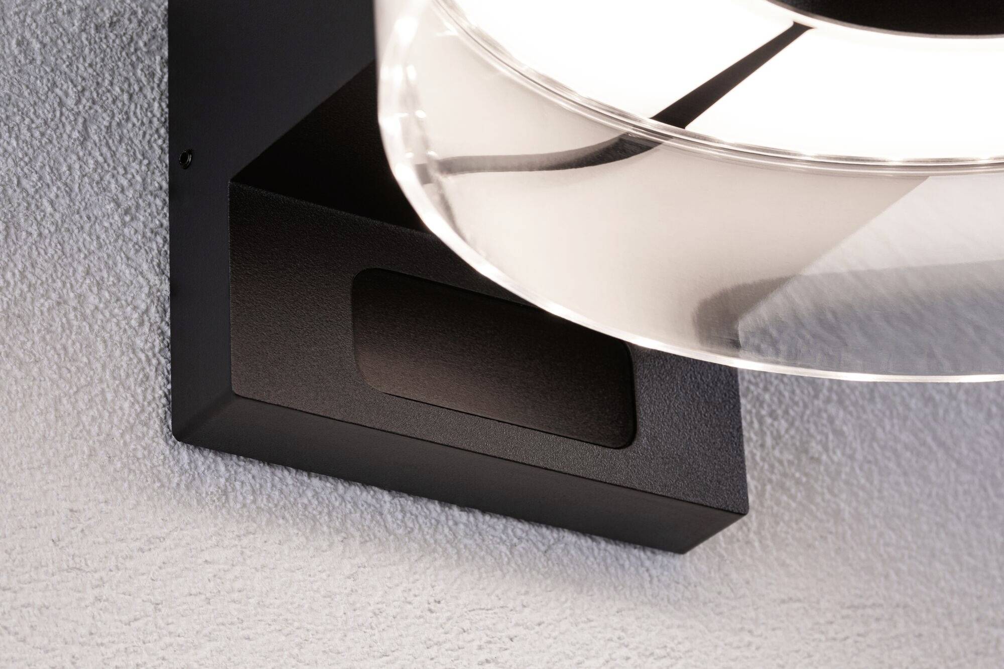 Eine moderne Wandlampe mit schwarzem Gehäuse und transparentem Schirm, die ein starkes Licht an eine strukturierte Wand wirft.