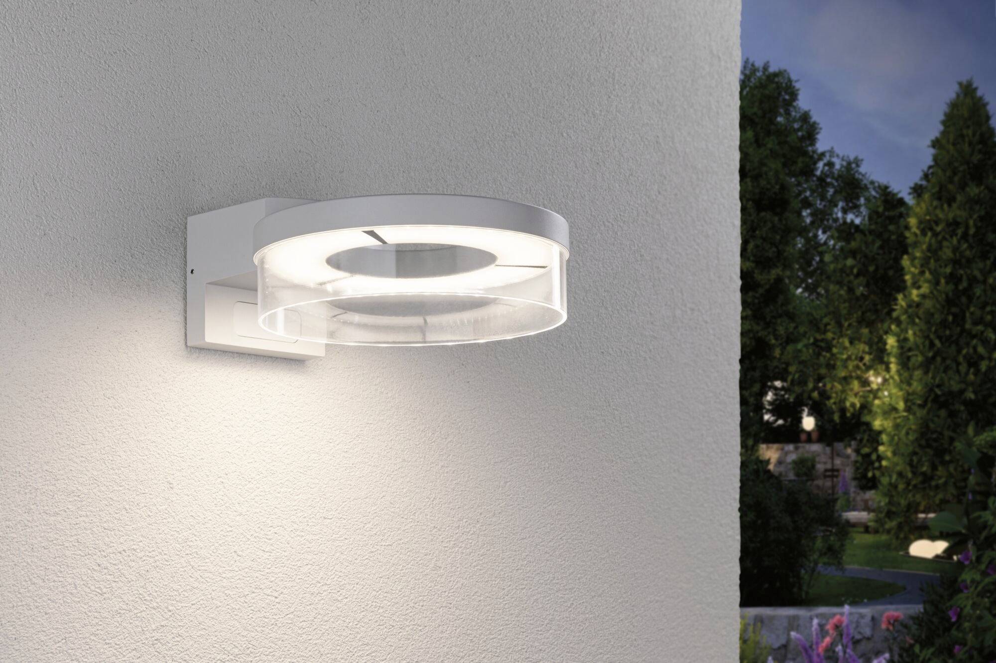 Moderne, runde Außenleuchte aus Metall und Glas, montiert an einer Wand. Beleuchtet eine Gartenfläche in abendlicher Umgebung.