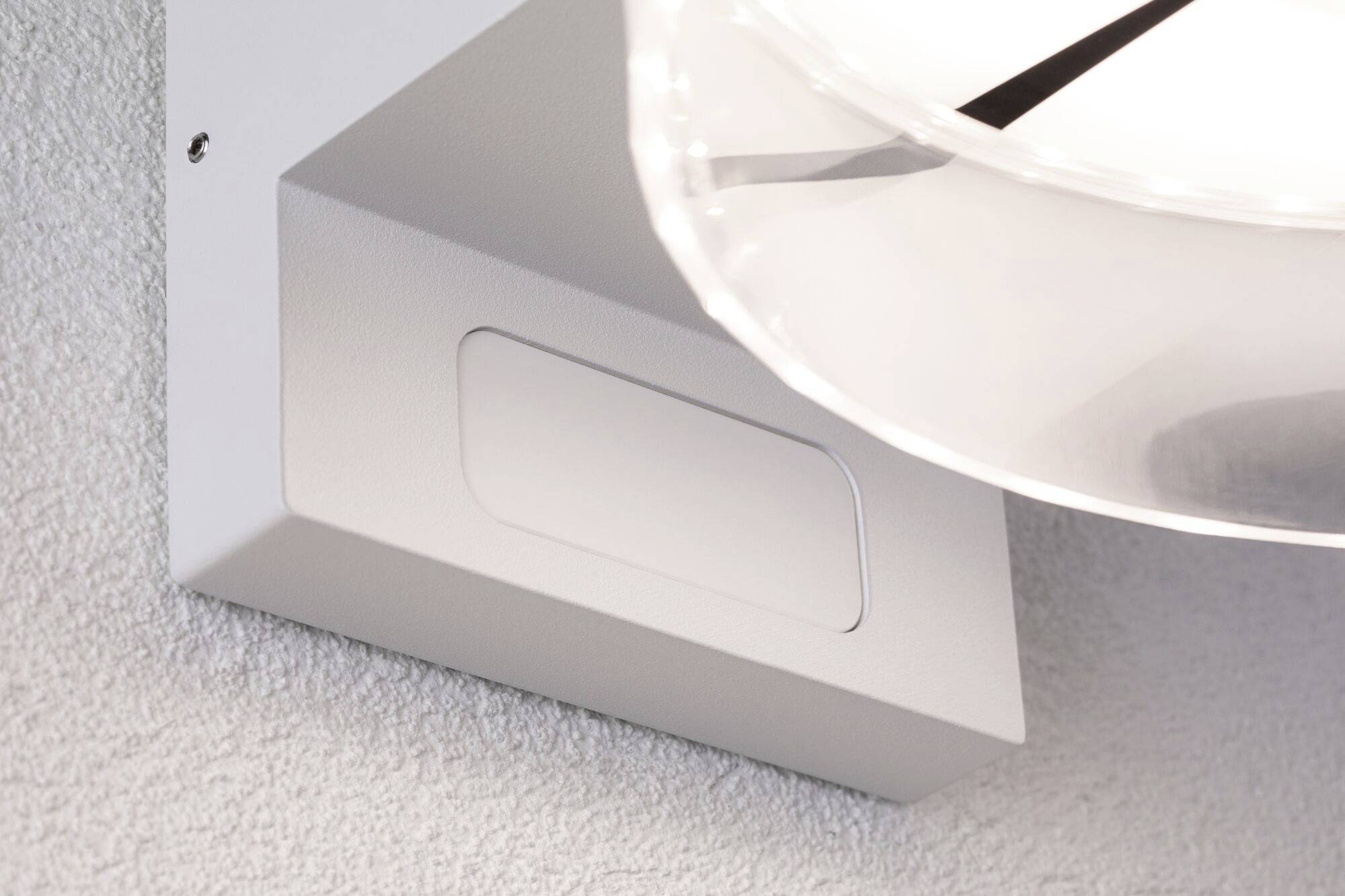 Eine weiße Wandleuchte mit moderner, minimalistischer Form, die sanftes Licht im Raum verteilt. Sie ist an einer strukturierten Wand montiert.