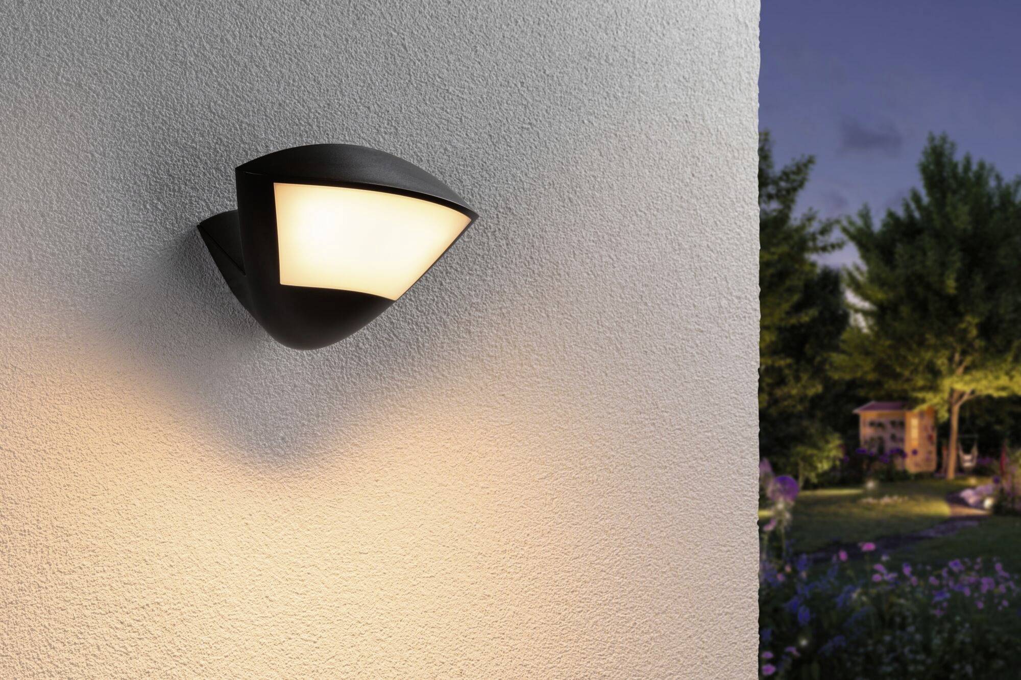 Eine moderne Außenwandleuchte leuchtet an einer weißen Wand, während im Hintergrund Bäume und ein Gartenhaus in der Abenddämmerung sichtbar sind.