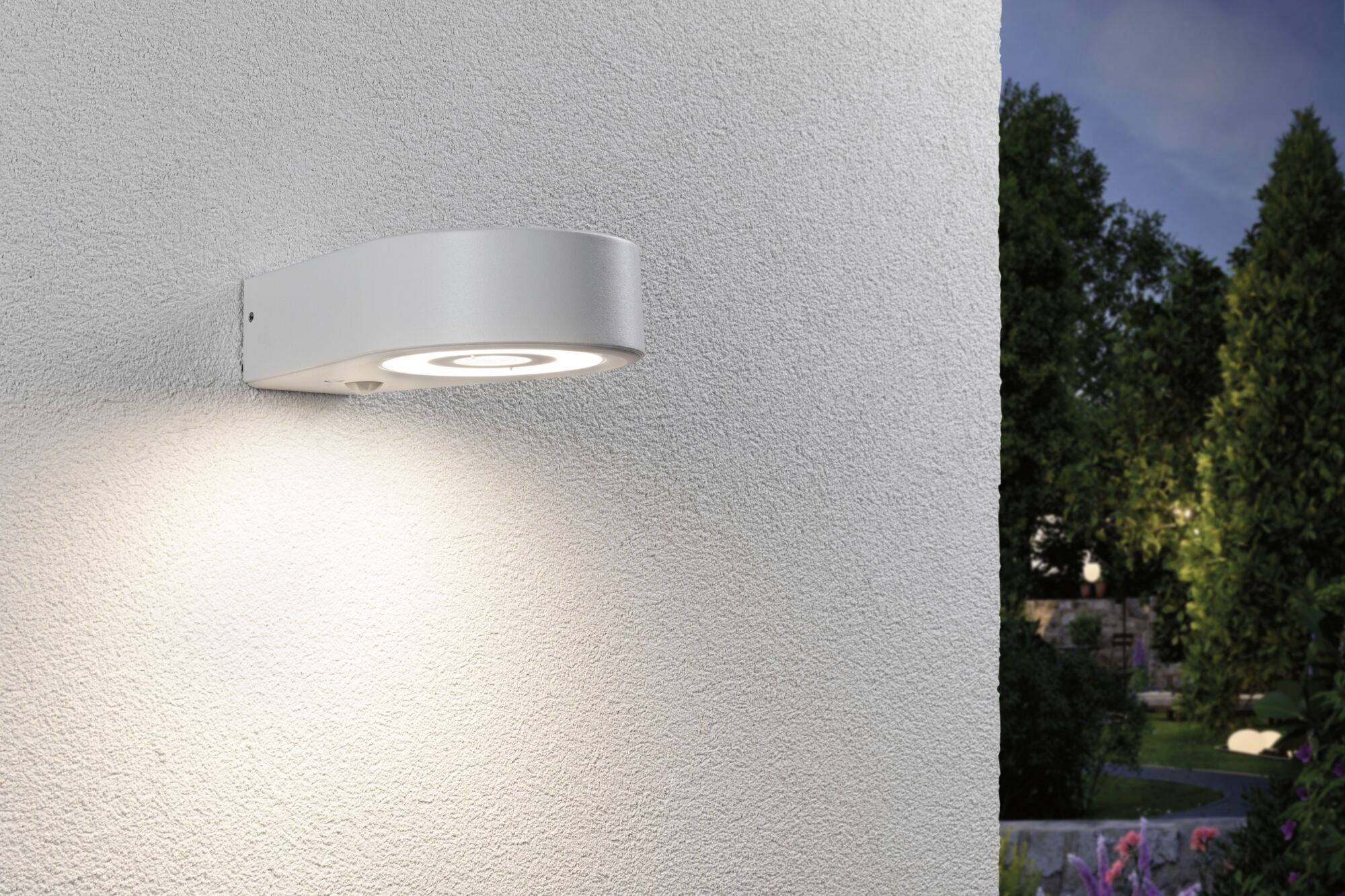 Moderne, runde Außenlampe an einer weißen Wand montiert, strahlt warmes Licht ab. Im Hintergrund sind Bäume und ein Garten bei Dämmerung.