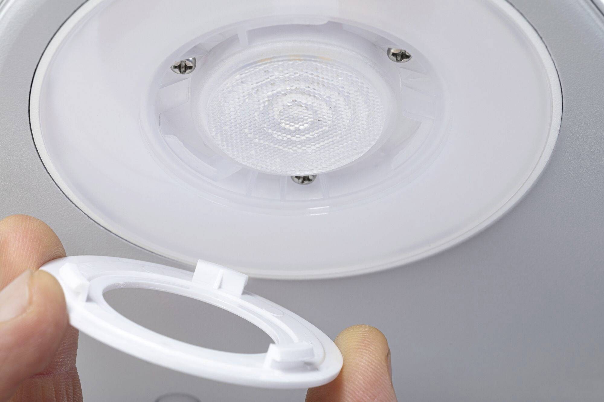 Eine Person hält einen runden Kunststoffrahmen und installiert ihn in eine Deckenleuchte, die eine kreisförmige LED-Lampe enthält.