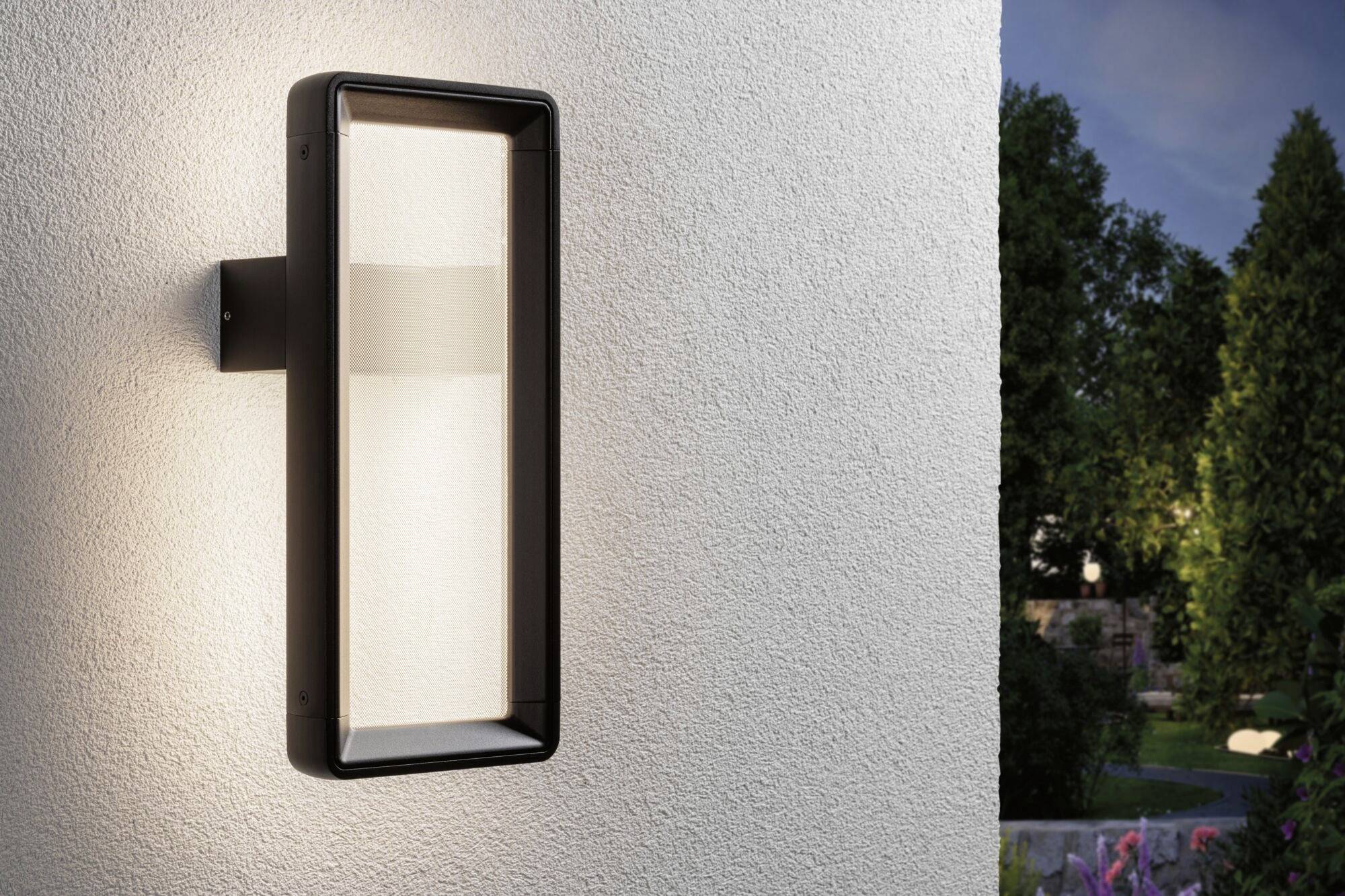 Eine moderne Außenleuchte an einer weißen Wand, beleuchtet einen Gartenbereich im Hintergrund bei Dämmerung.