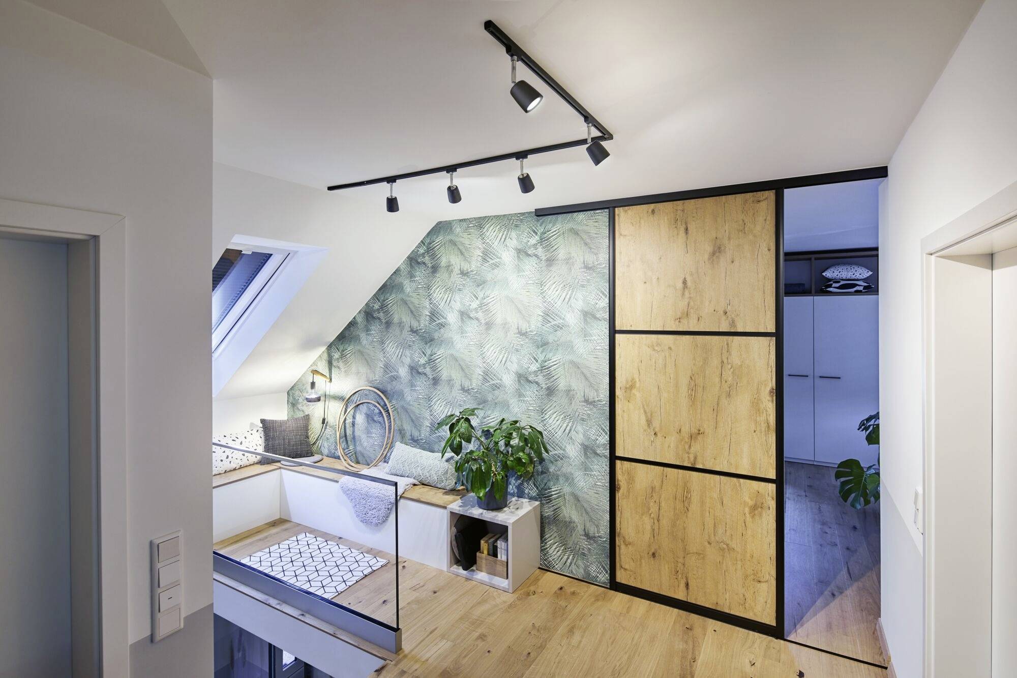 Ein helles Dachgeschosszimmer mit Holzfußboden, einer botanisch gemusterten Wand und einem eingebauten Schrank. Eine Pflanze steht neben einer Sitznische.