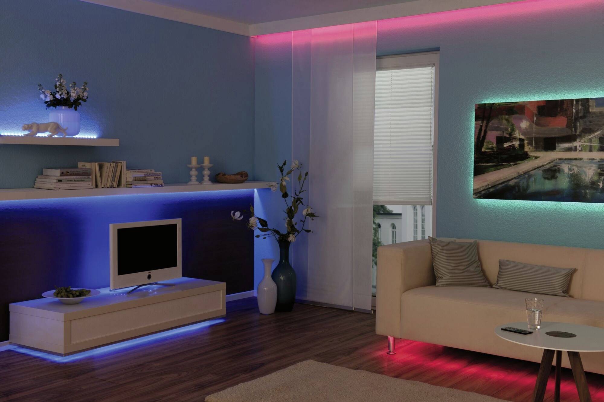 Wohnzimmer mit blauer LED-Beleuchtung unter Regal, roter Beleuchtung an Decke, Bild mit grüner Beleuchtung, Sofa und Fernseher.