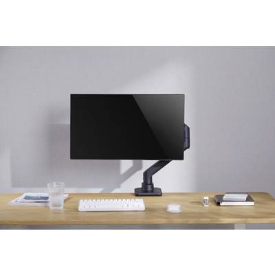 Ein Schreibtisch mit Monitor auf Ständer, daneben Tastatur, Maus, Glas, Fernbedienung, Notizbuch und Zeitschrift, vor grauer Wand.