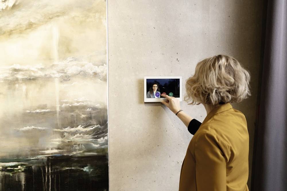 Frau mit blondem Haar und gelbem Blazer nutzt an einer Wand ein Tablet für einen Videoanruf, daneben abstrakte Kunst.