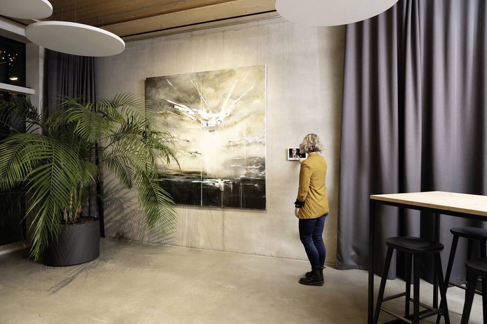 Eine Person fotografiert ein großes Gemälde an einer Wand. Der Raum ist modern mit Pflanzen, Vorhängen und einem hohen Tisch mit Barhockern.