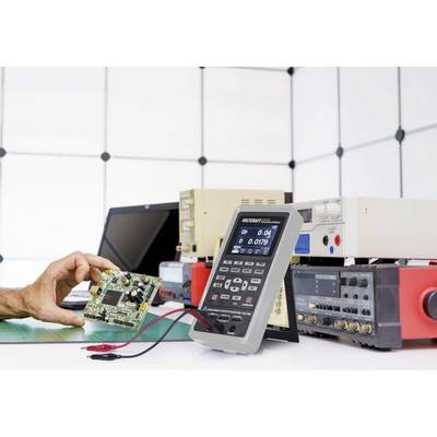 Eine Person hält eine Computerplatine neben mehreren elektronischen Messgeräten auf einem Tisch. Es handelt sich um ein Laborumfeld.