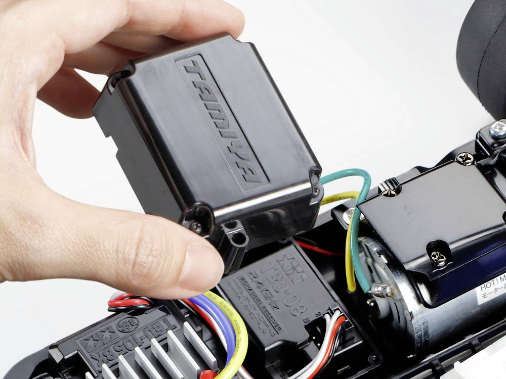 Eine Hand montiert ein schwarzes elektronisches Bauteil mit der Aufschrift 'TRU....' auf eine Platine eines ferngesteuerten Autos.