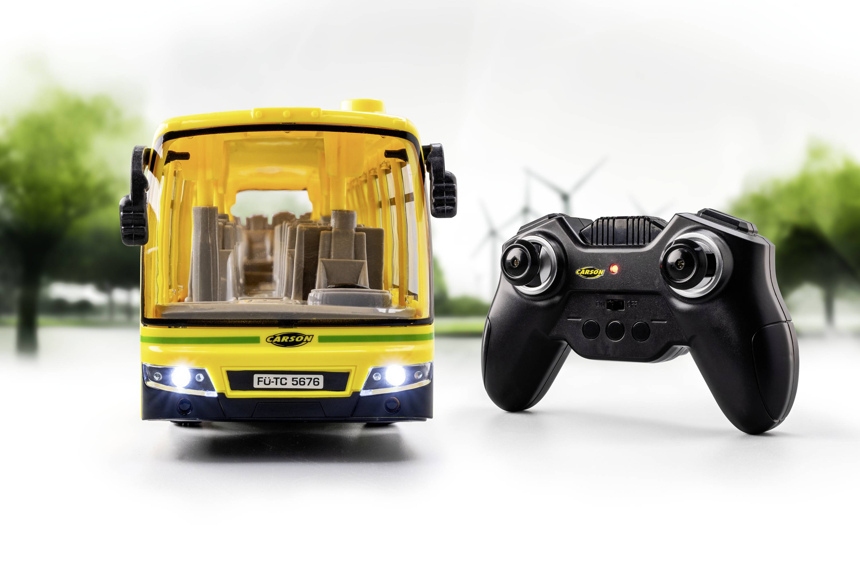 Spielzeugbus mit gelb-schwarzem Design und Fernsteuerung auf unscharfem Hintergrund mit Bäumen und Windrädern.