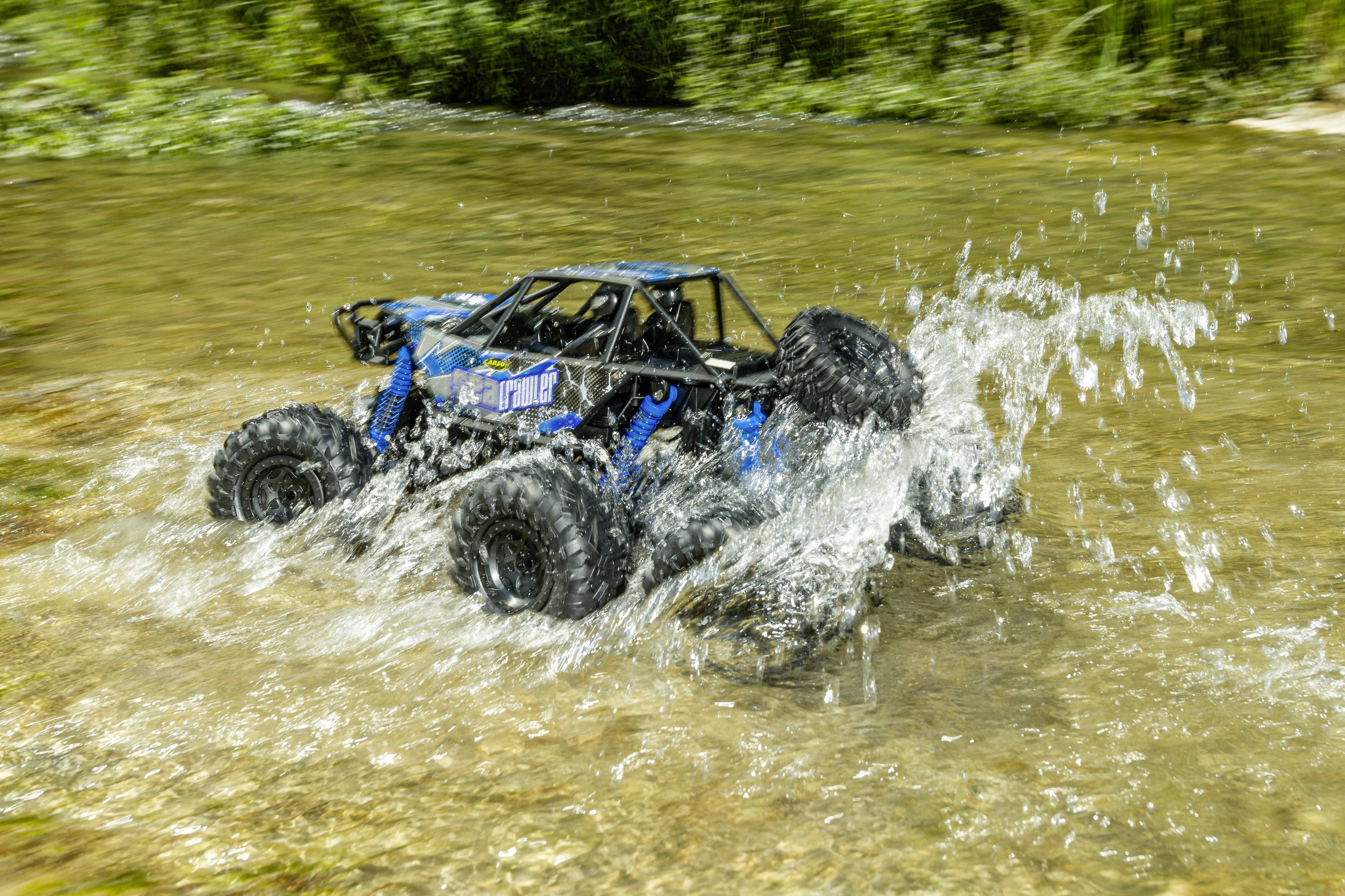 Ein ferngesteuertes Auto fährt durch flaches Wasser und spritzt dabei Wasser auf. Es befindet sich in einer natürlichen Umgebung.
