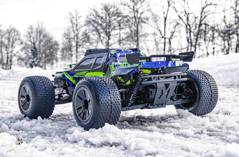 Ein ferngesteuertes Auto fährt auf schneebedecktem Boden. Es hat große Reifen und ein buntes, auffälliges Design. Im Hintergrund sind Bäume.