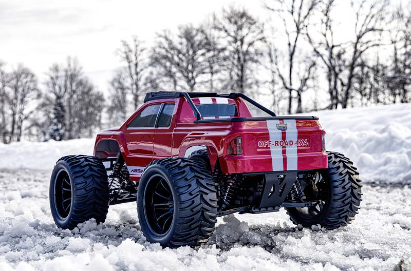 Ein roter Offroad-Truck mit großen Reifen steht im verschneiten Gelände. Im Hintergrund sind kahlen Bäume und ein bewölkter Himmel.