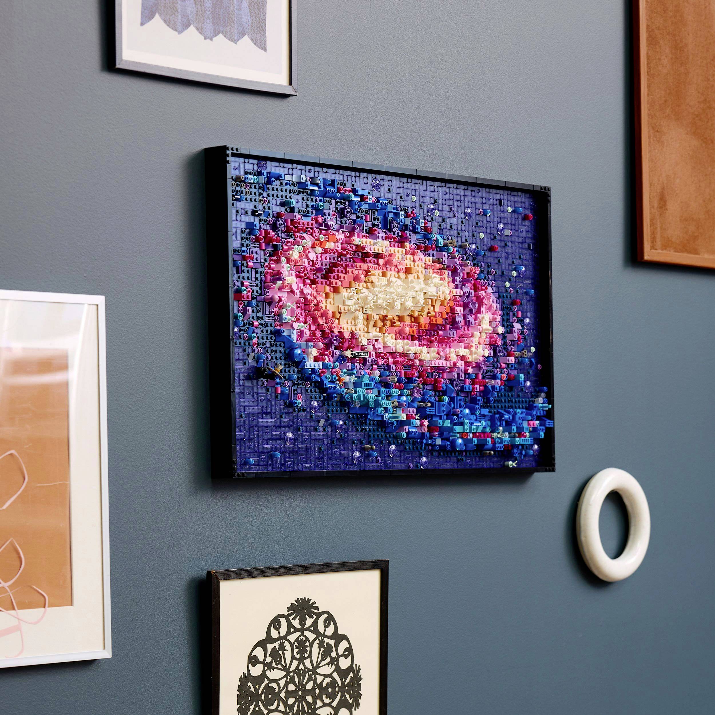 Mehrere abstrakte Kunstwerke an einer blauen Wand, darunter ein farbenfrohes Mosaik, das eine Galaxie darstellt.