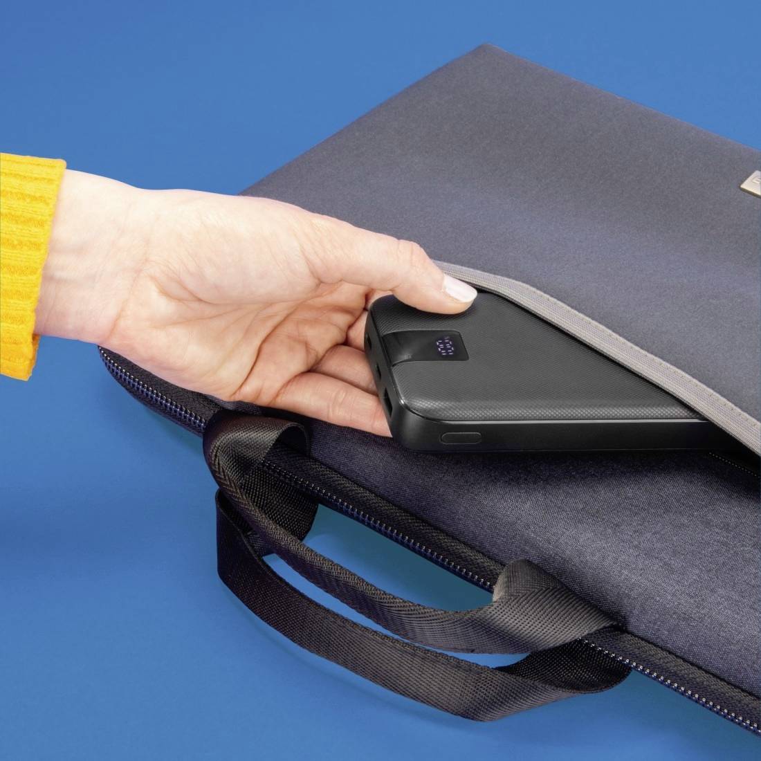 Eine Hand hält ein schwarzes, tragbares Ladegerät über einer grauen Tasche auf einem blauen Hintergrund.