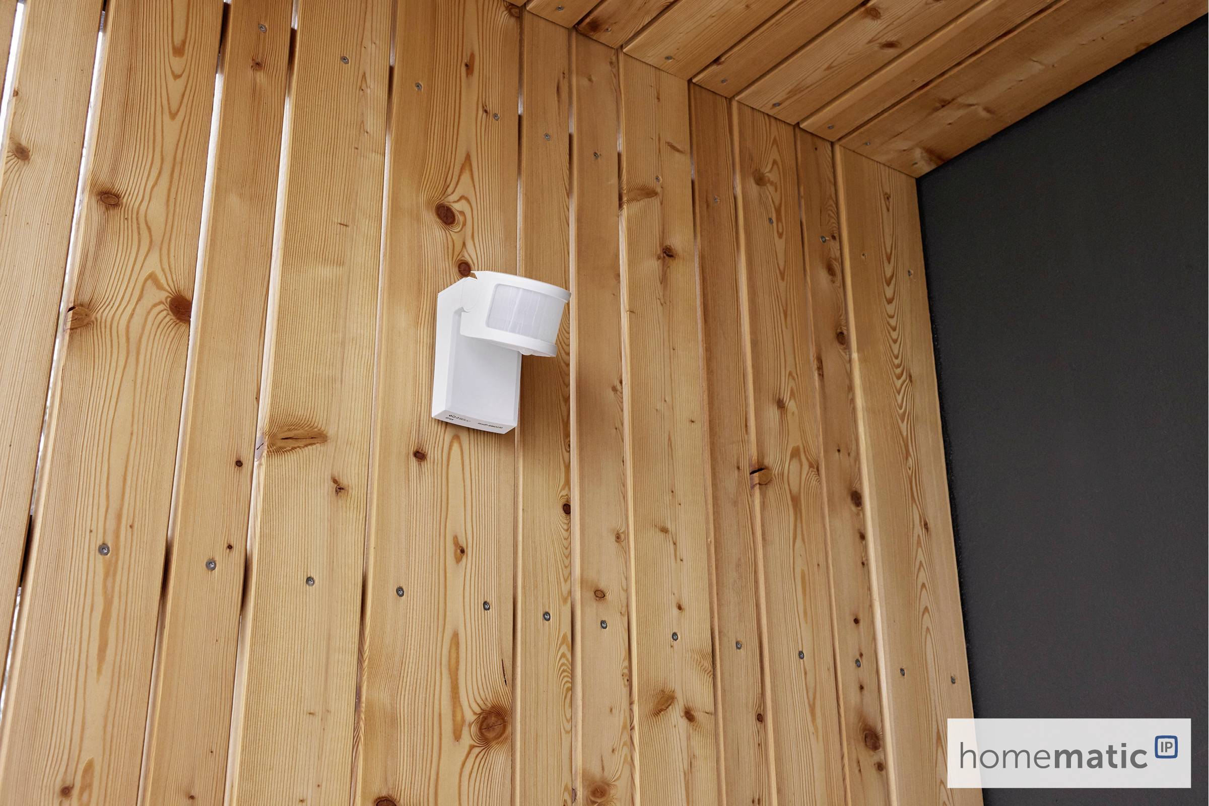 An einer hölzernen Wand ist ein weißer Bewegungsmelder montiert. Unten rechts im Bild befindet sich ein 'homematic'-Logo.