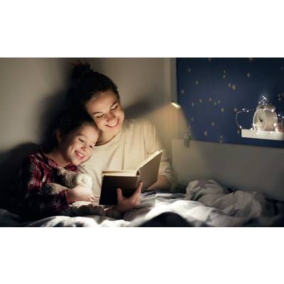 Frau und Kind sitzen im Bett und lesen ein Buch bei warmem Licht. Das Kind kuschelt mit einem Stofftier. Im Hintergrund Sterne.