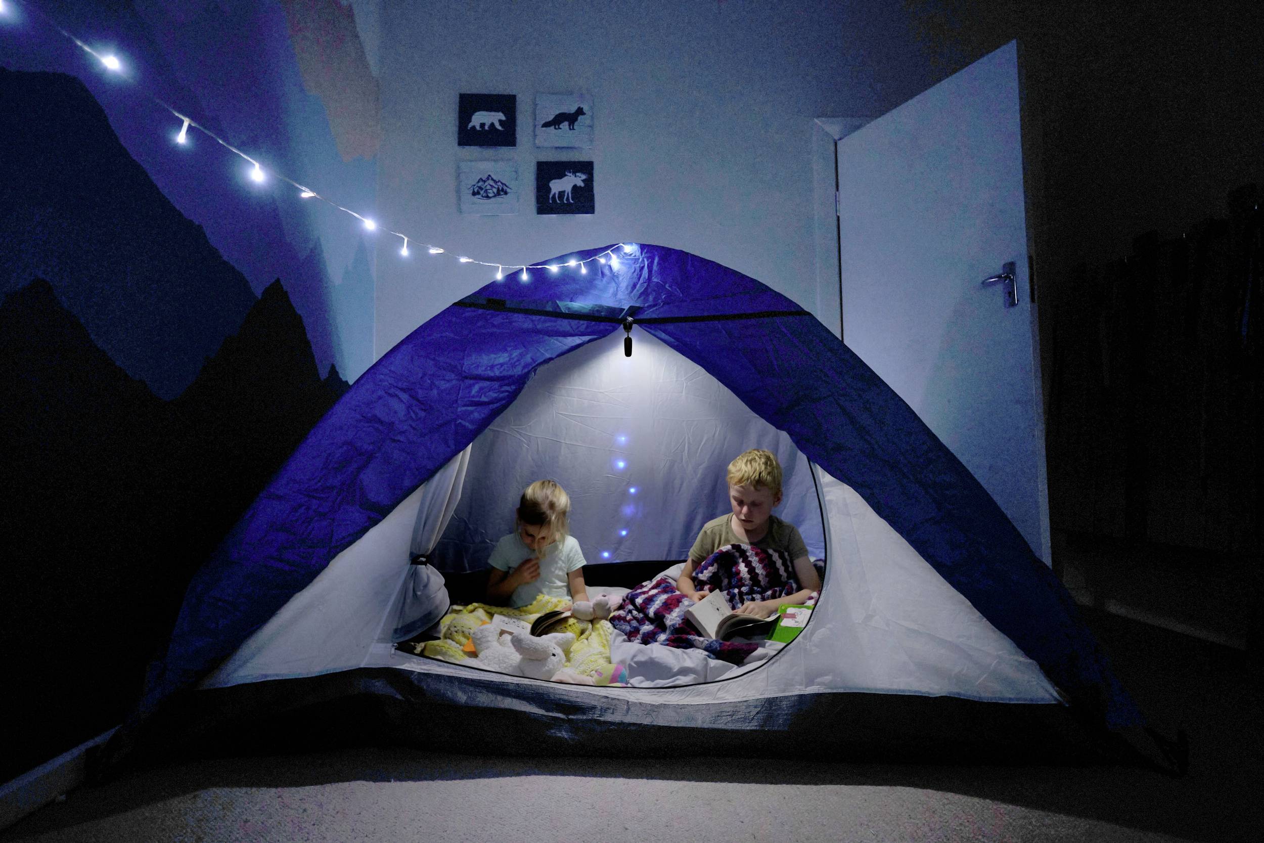 Zwei Kinder sitzen in einem Zelt in einem dunklen Zimmer, umgeben von Lichterketten. Sie lesen und wirken konzentriert.