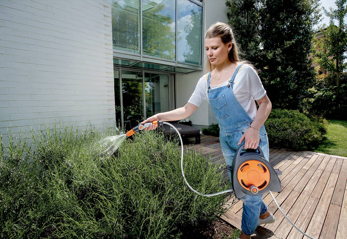 Eine Frau gießt mit einem Schlauch Pflanzen im Garten vor einem modernen Gebäude. Sie trägt einen weißen T-Shirt und blaue Latzhose.