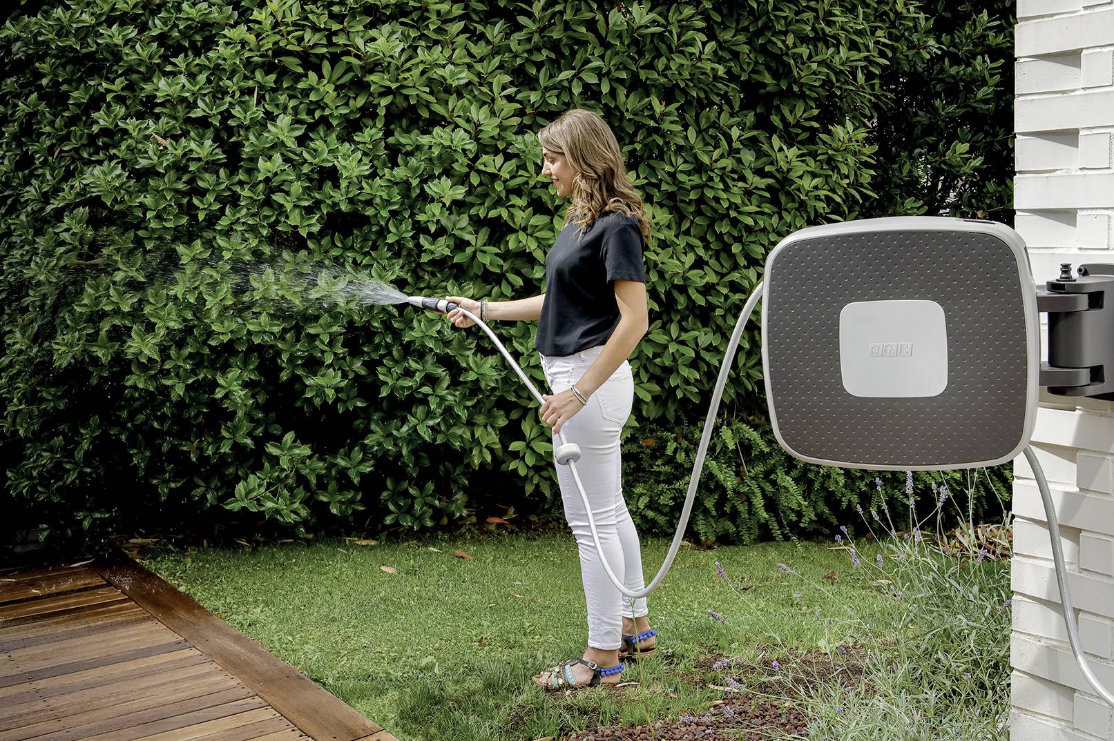 'Frau gießt mit einem Wasserschlauch den Rasen in einem Garten. Im Hintergrund sind dichte grüne Büsche zu sehen.'