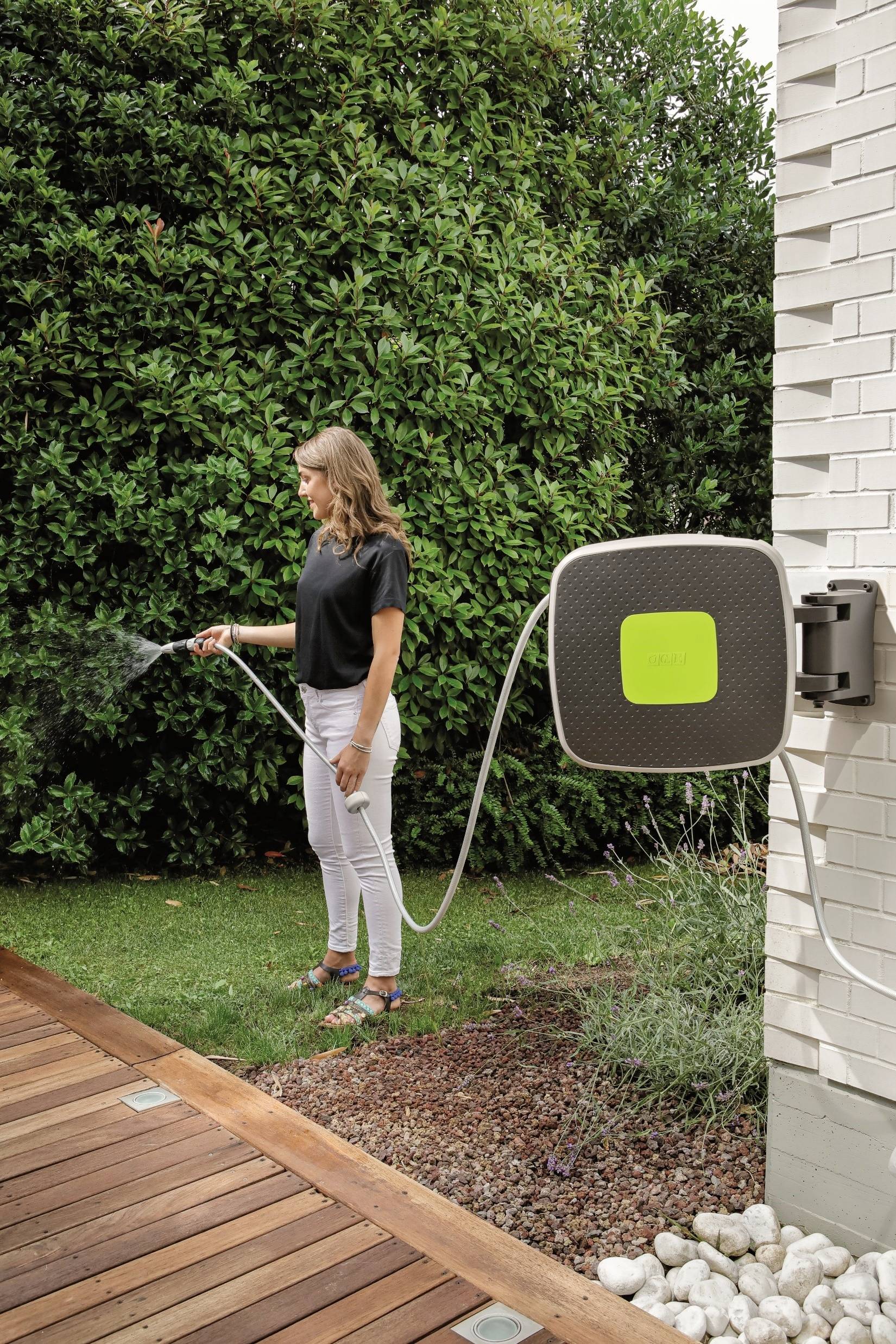 Frau gießt den Garten mit einem Schlauch, der an einer weißen Wand befestigt ist. Im Hintergrund sind grüne Büsche zu sehen.