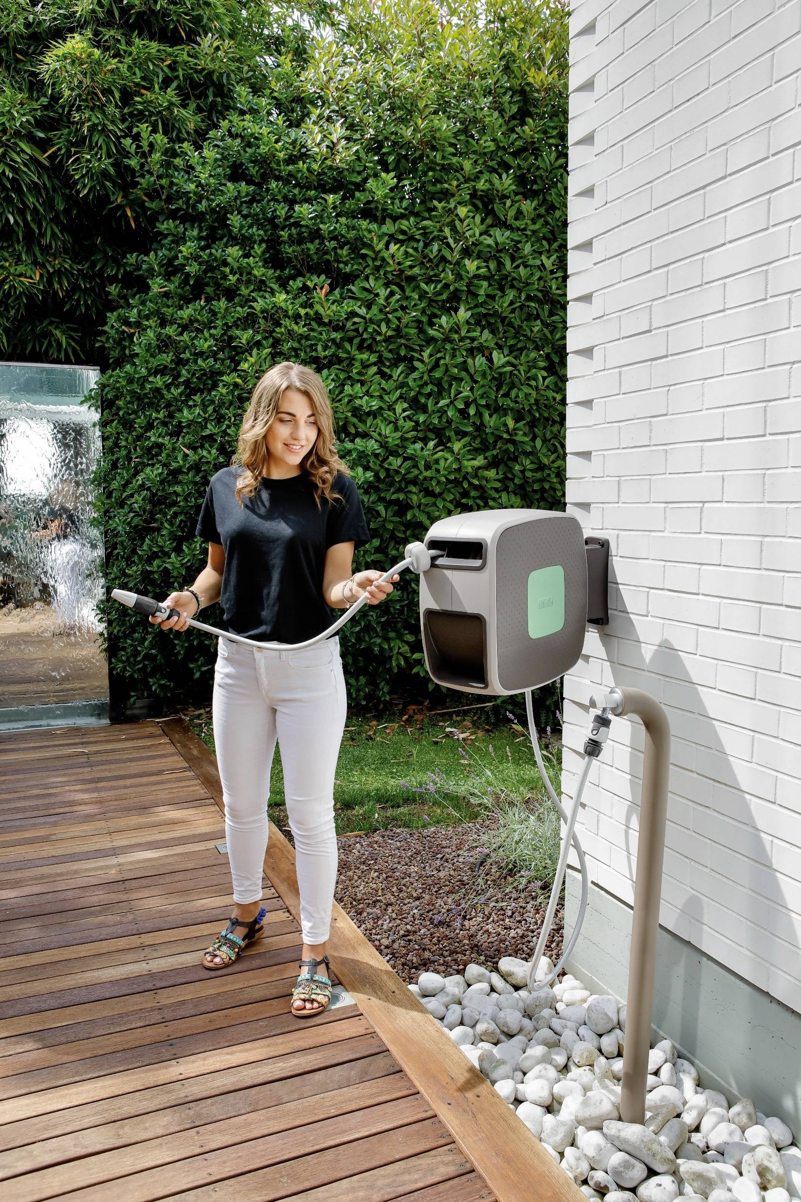 Eine Frau in Freizeitkleidung steht auf einer Holzterrasse vor einem weißen Haus und hält ein Ladekabel für ein Elektroauto.