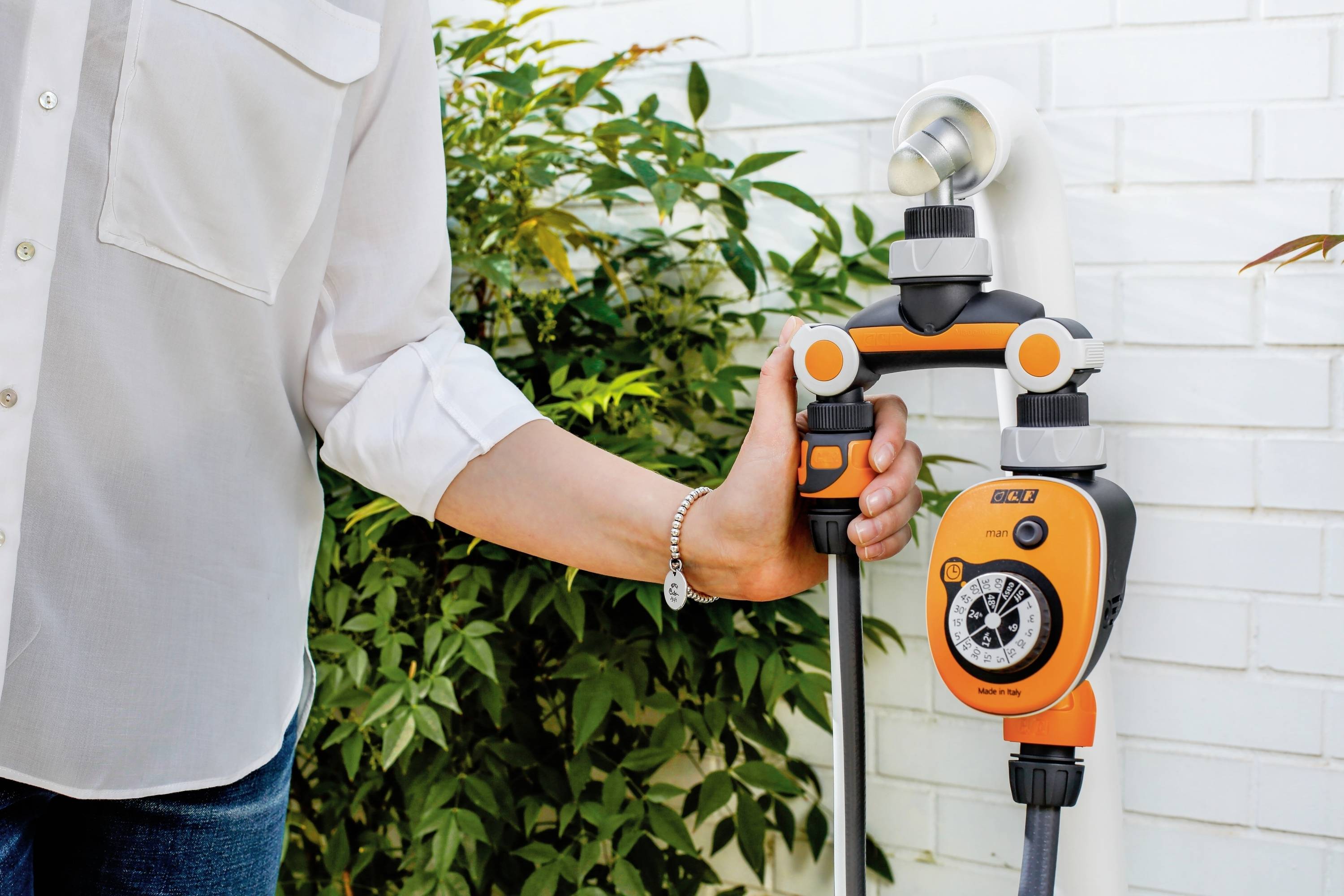 Eine Person installiert einen orange-schwarzen Bewässerungstimer an einem Wasserhahn im Garten, umgeben von Pflanzen und einer weißen Wand.