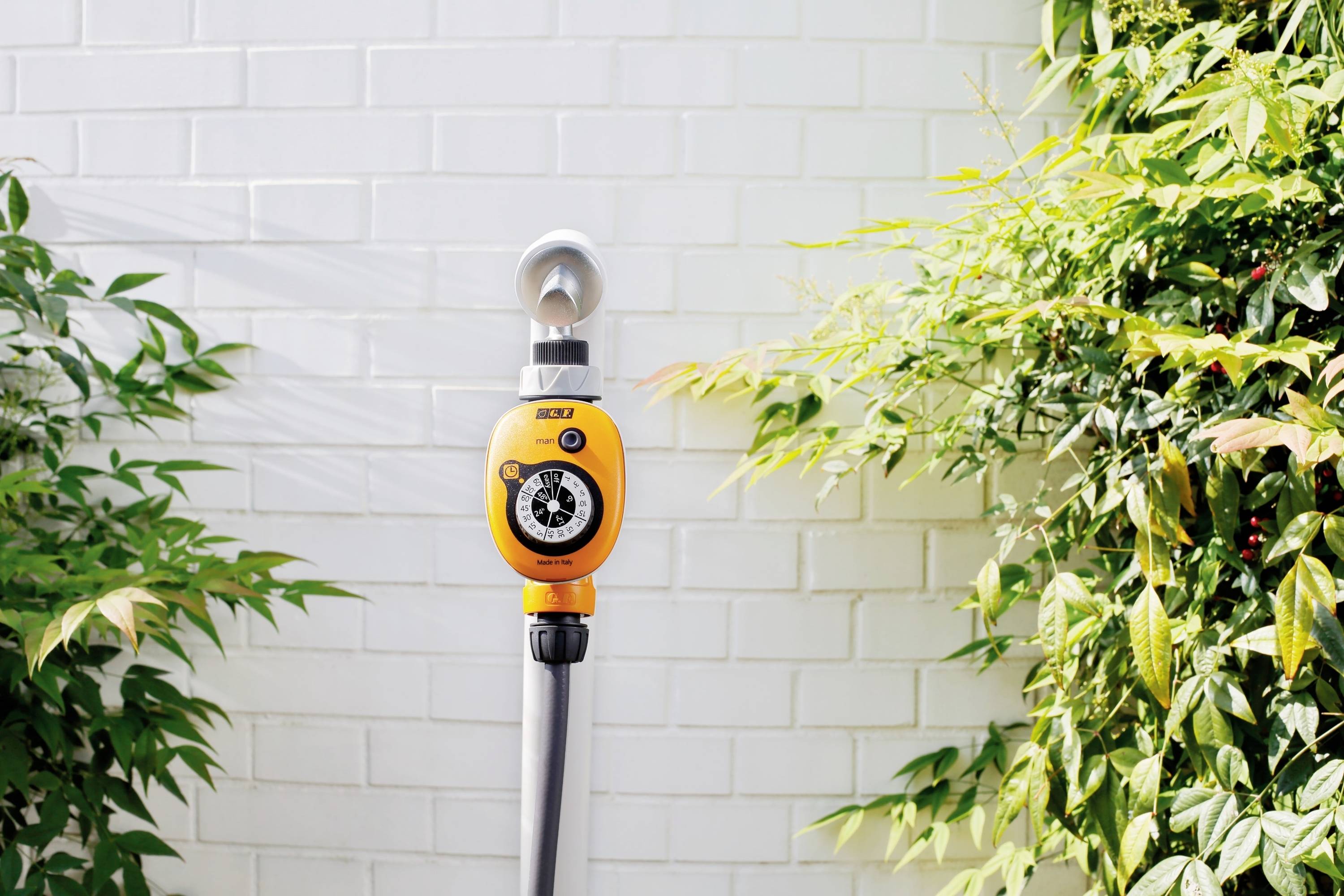 'Grauer Gartenschlauch mit gelbem Wasserhahn-Timer vor einer weißen Ziegelwand, umgeben von grünen Pflanzen.'