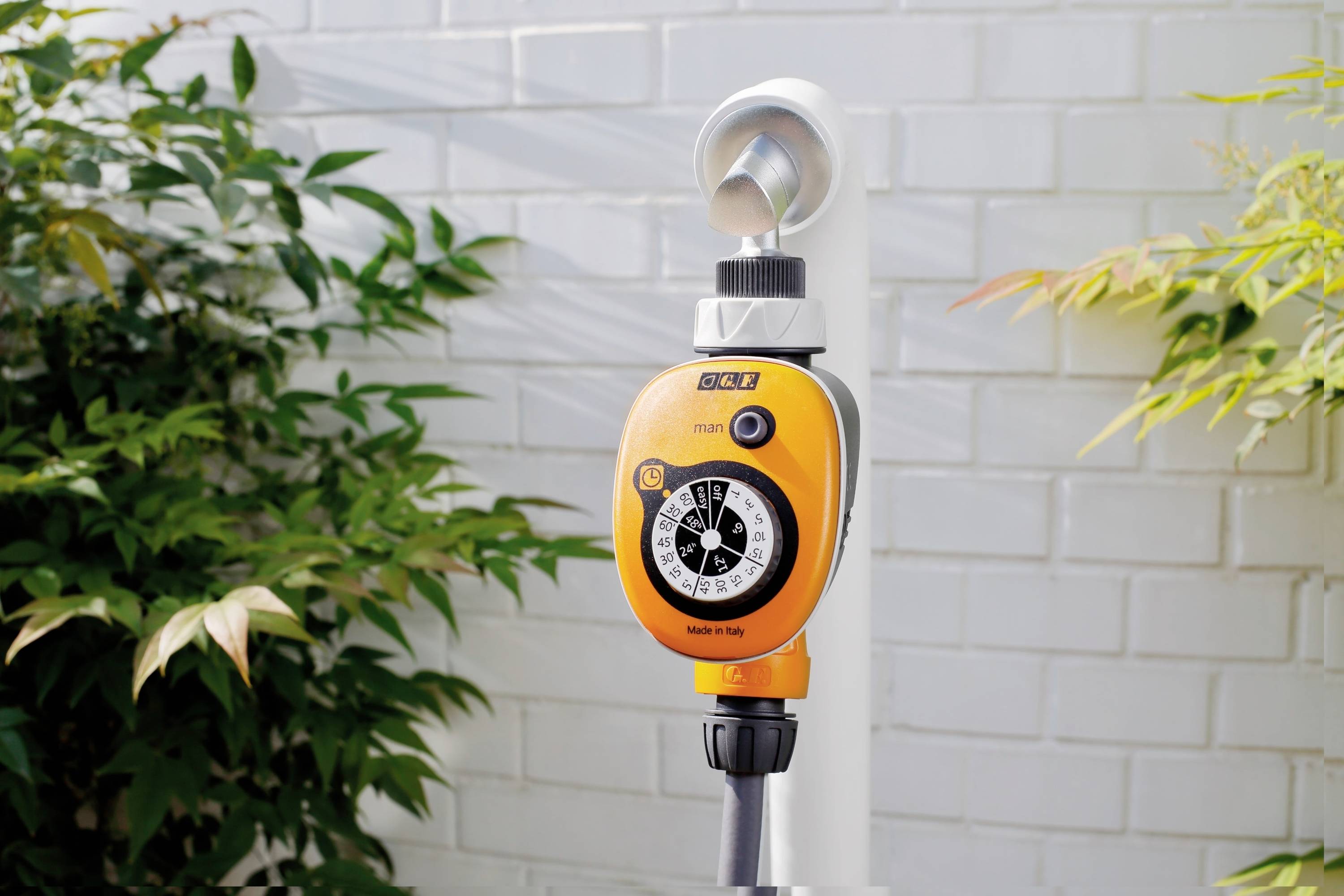 Ein orangefarbener Garten-Bewässerungs-Timer ist an einem weißen Rohr befestigt. Im Hintergrund sind Ziegelwände und Pflanzen zu sehen.