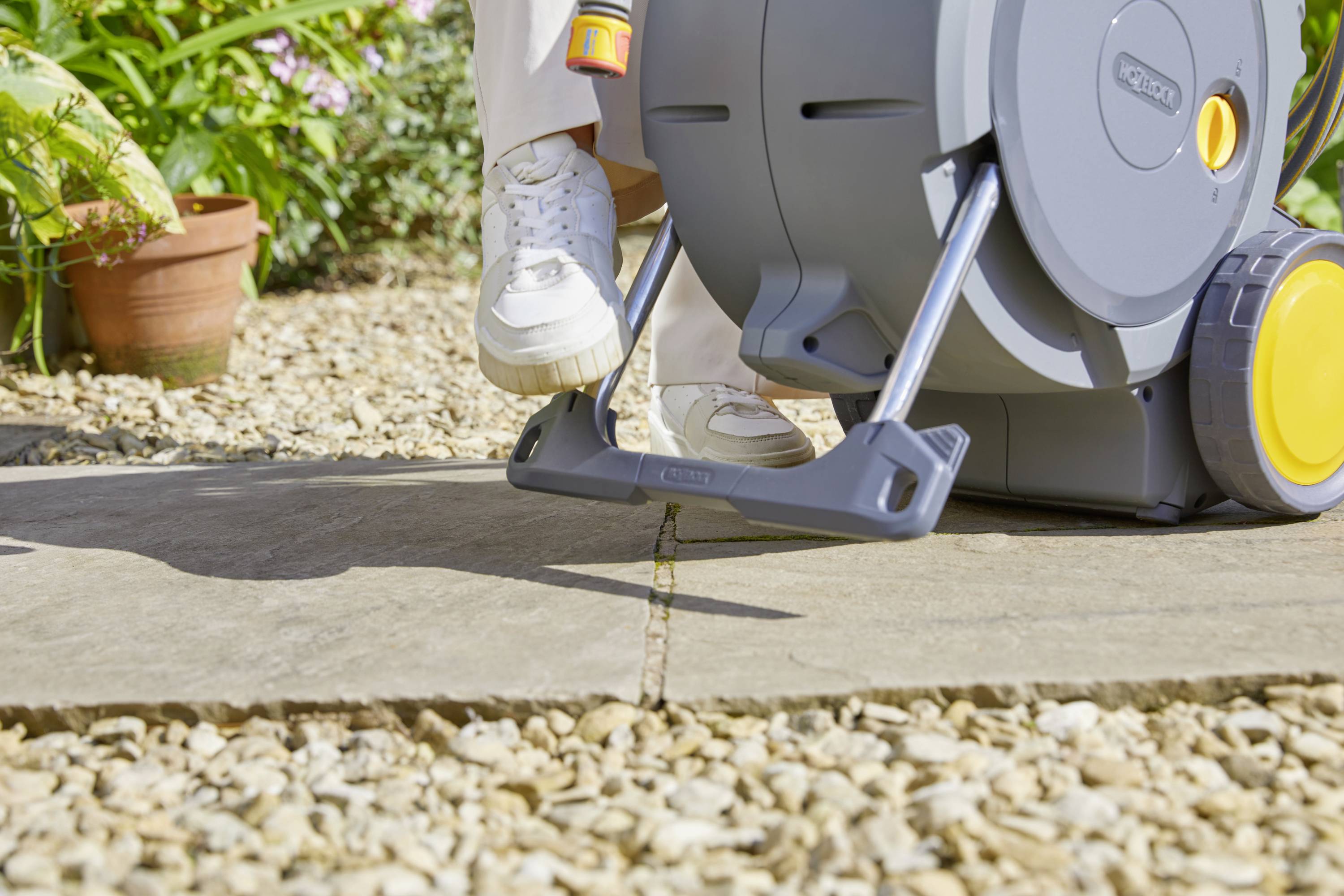 Eine Person drückt mit dem Fuß auf einen Schlauchwagen im Garten. Der Schlauchwagen hat Räder und steht auf Steinplatten.